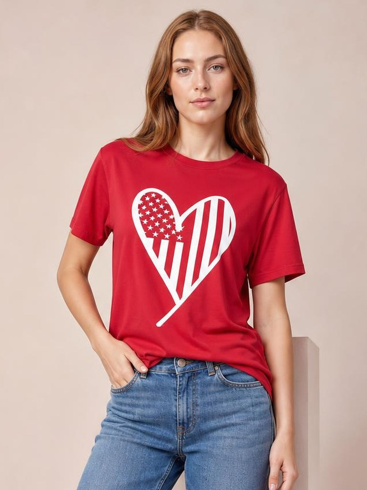 A woman wears a red round neck short sleeve T-shirt with a bold white heart graphic, relaxed basic silhouette, and one hand tucked into her jeans pocket.