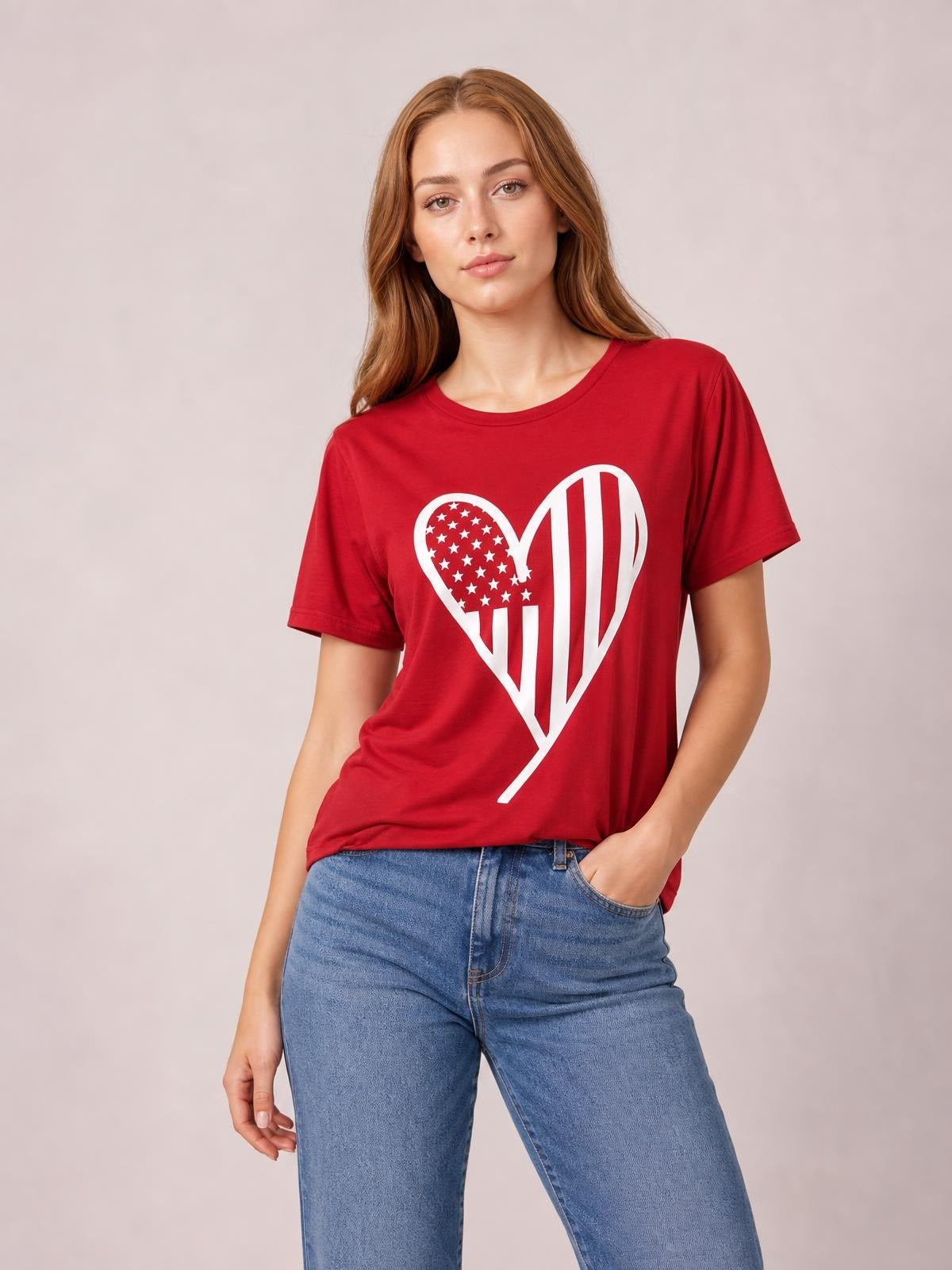 Model wearing a red heart round neck short sleeve T-shirt with a white patriotic heart graphic, relaxed silhouette, and one hand in her jeans pocket.