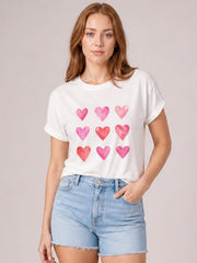 A model poses in a white round-neck short-sleeve T-shirt with rolled cuffs, a relaxed silhouette, and a playful grid of pink and red hearts.