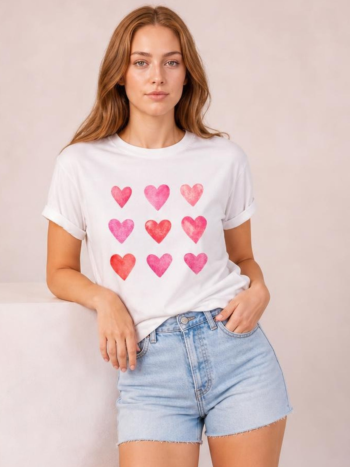 Model wearing a white Heart Round Neck Short Sleeve T-Shirt with rolled cuffs and nine colorful heart prints, shown in a relaxed pose with one hand in her shorts pocket.