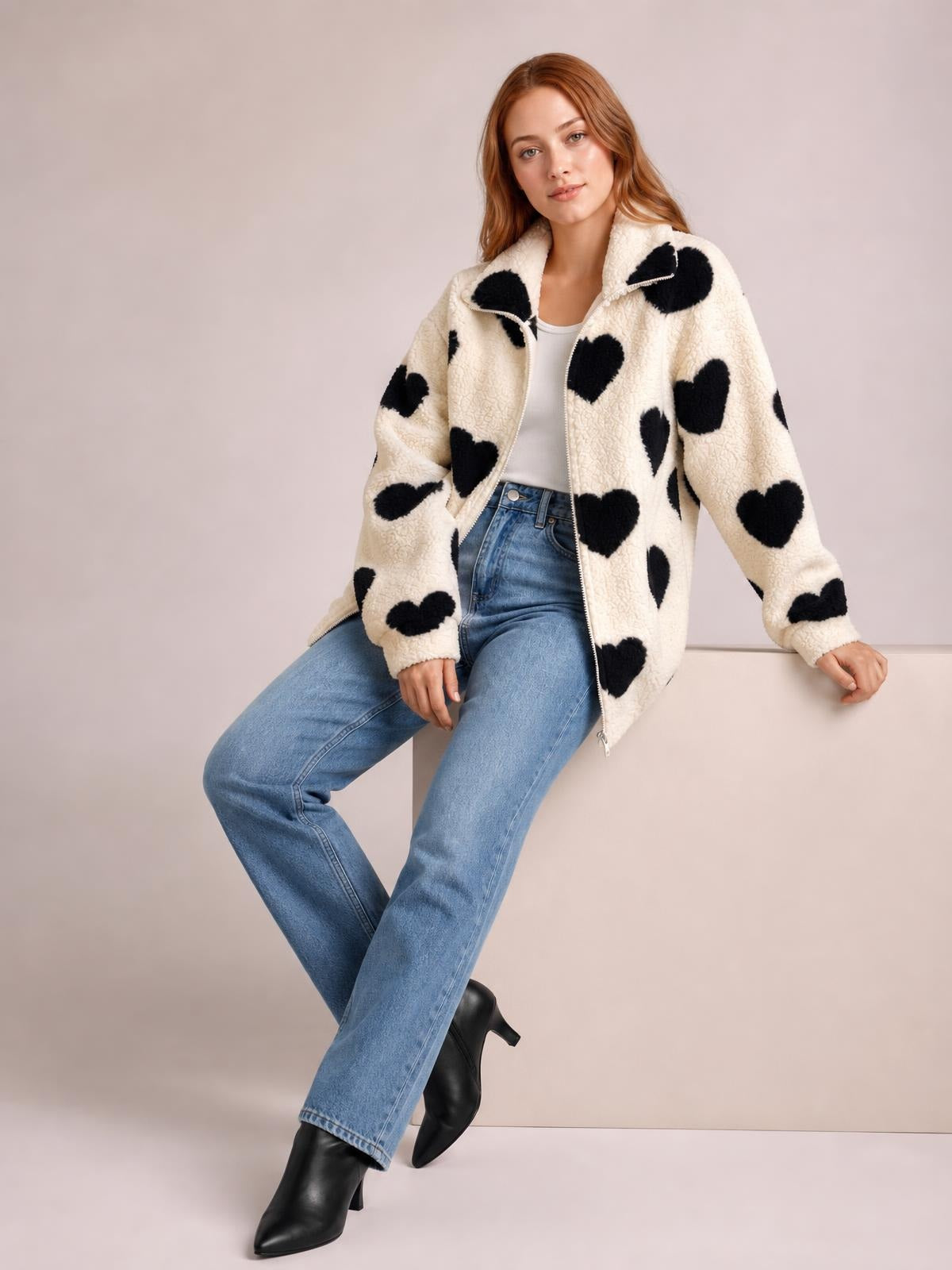 A model sits in a cream furry heart-print jacket with a front zip, drop shoulders, relaxed silhouette, and long sleeves, paired with jeans.
