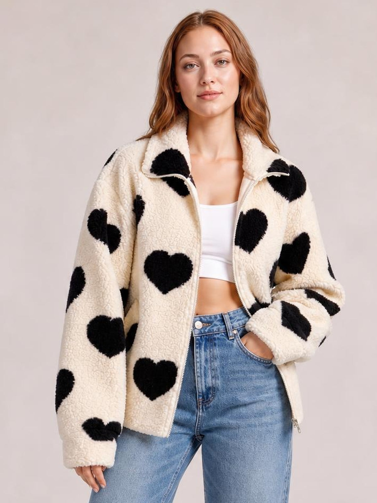A model poses in a cream furry drop-shoulder jacket with a front zip, oversized black heart motifs, and a relaxed silhouette, one hand in a pocket.