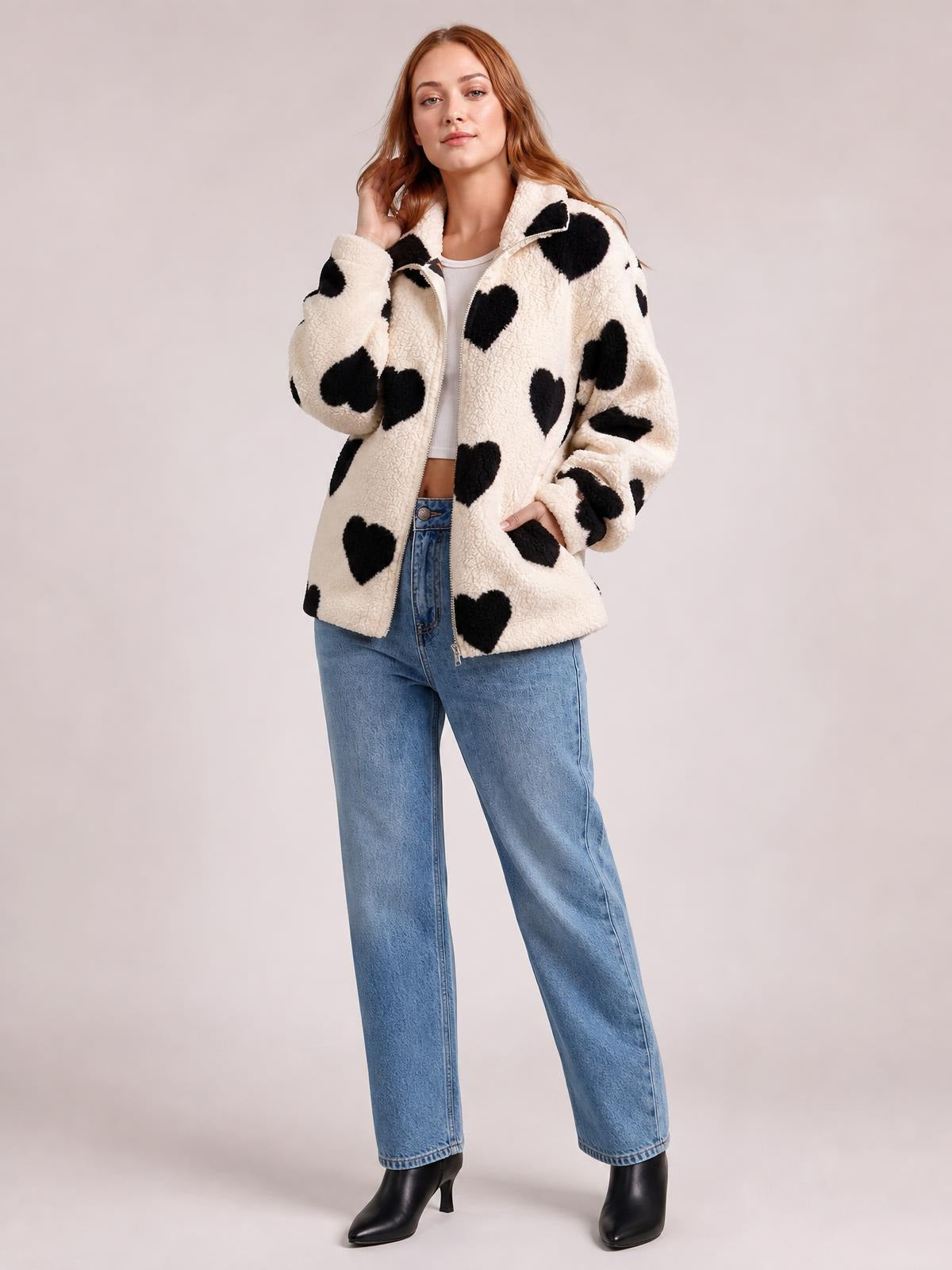 A model poses in a cream furry zip-up jacket with black heart prints, a relaxed drop-shoulder silhouette, stand collar, and long sleeves.