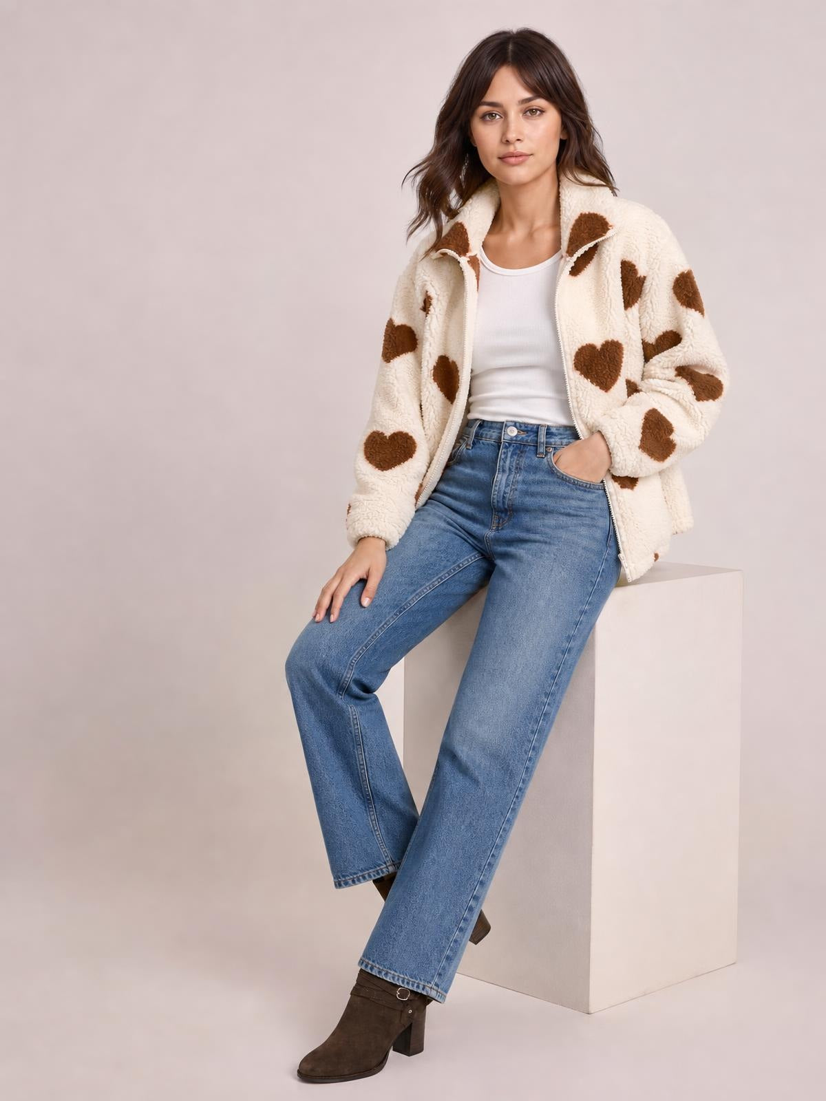 A model sits in a cream furry heart zip-up jacket with brown heart motifs, drop shoulders, a relaxed silhouette, and a front zipper.