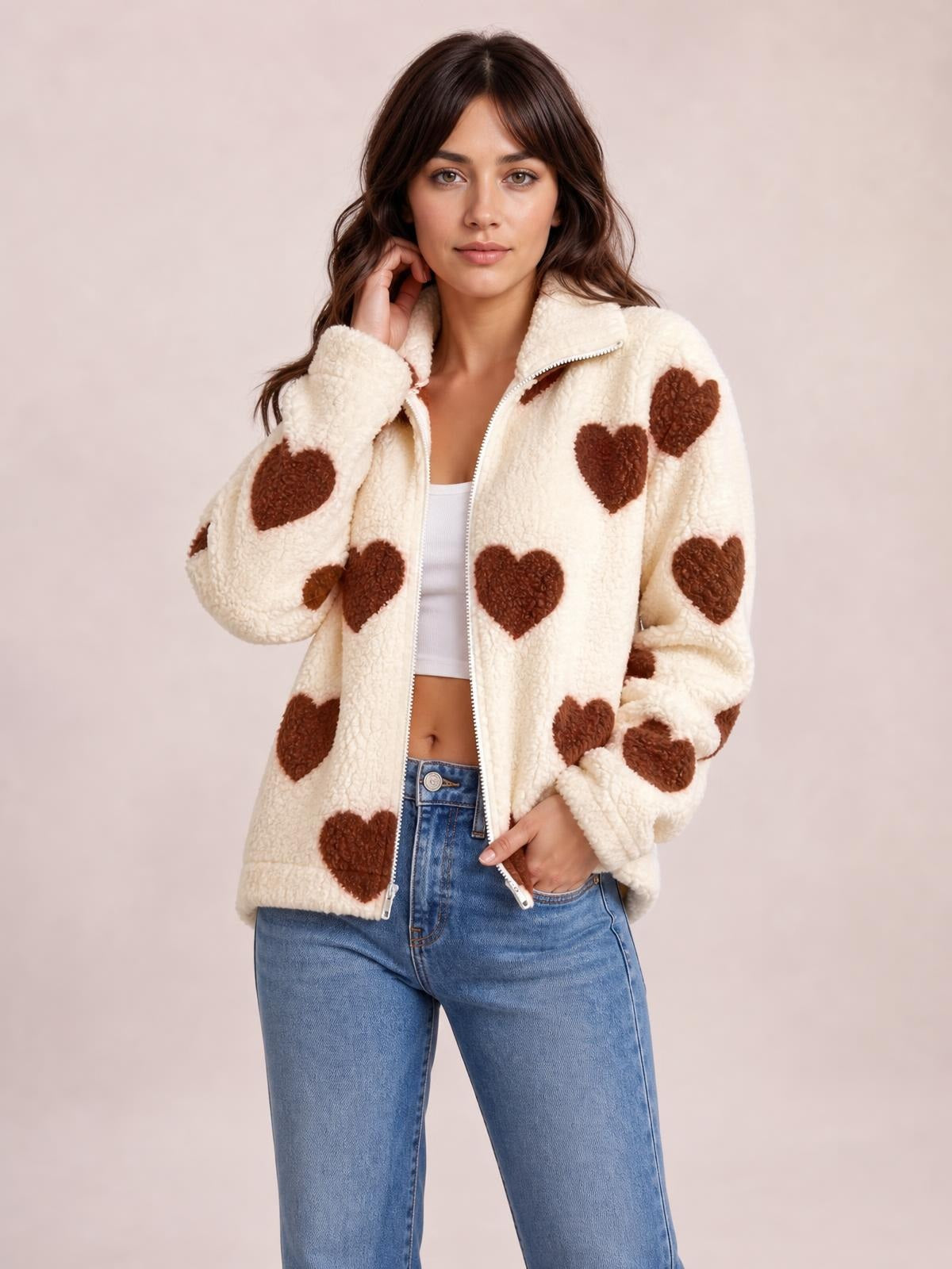 Model poses in a cream furry heart jacket with brown heart patches, drop shoulders, a zip front, stand collar, and relaxed silhouette.