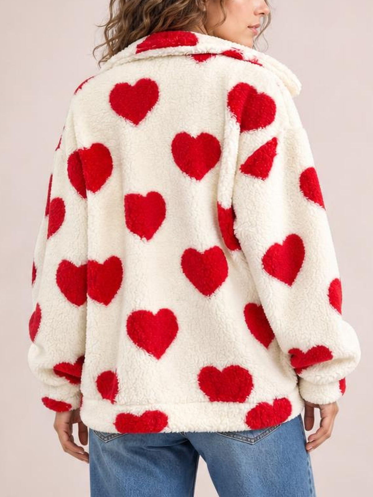 A model shows the back of a cream furry jacket with red heart pattern, hood, drop shoulders, relaxed silhouette, and hands resting at her sides.