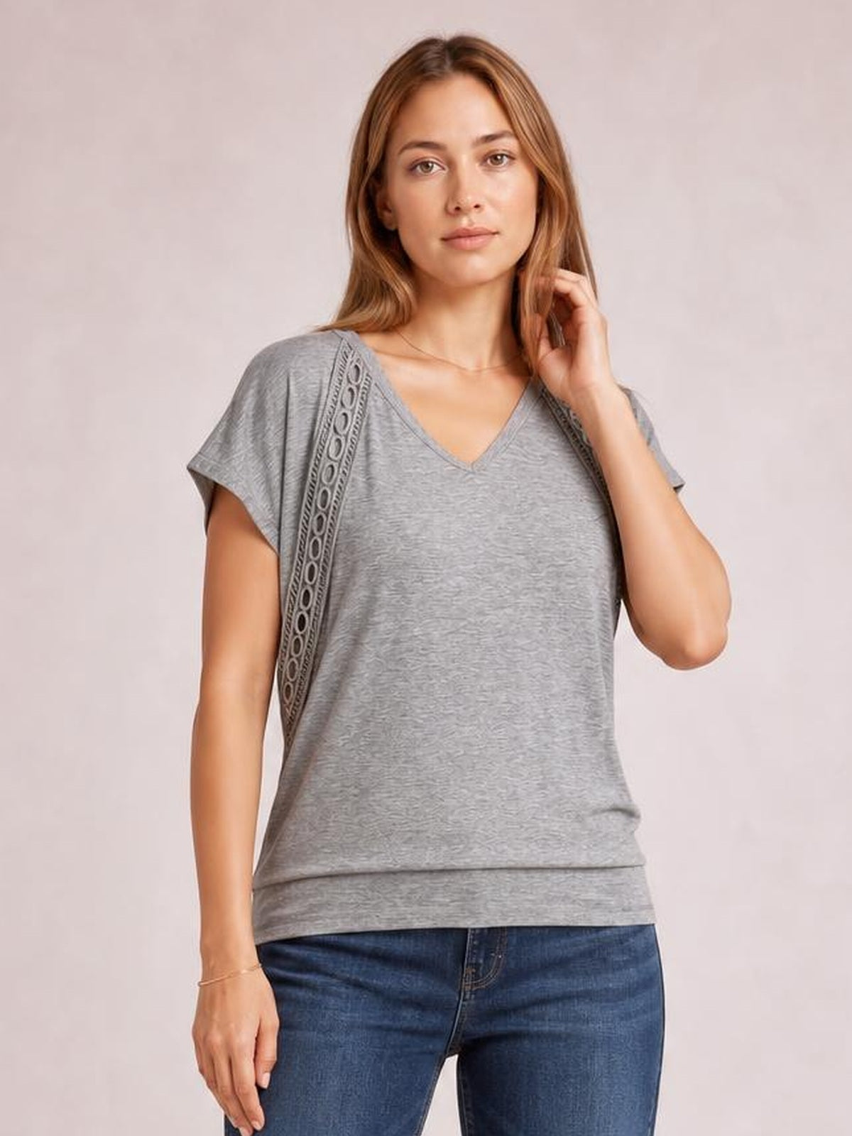 Model wears a gray heathered V-neck short sleeve T-shirt with a relaxed silhouette and eyelet-trimmed shoulder panels, posing with one hand in her hair.