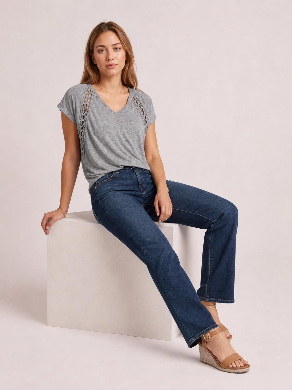 Model seated in a relaxed pose wearing a gray heathered V-neck short-sleeve T-shirt with eyelet shoulder details and a loose silhouette.