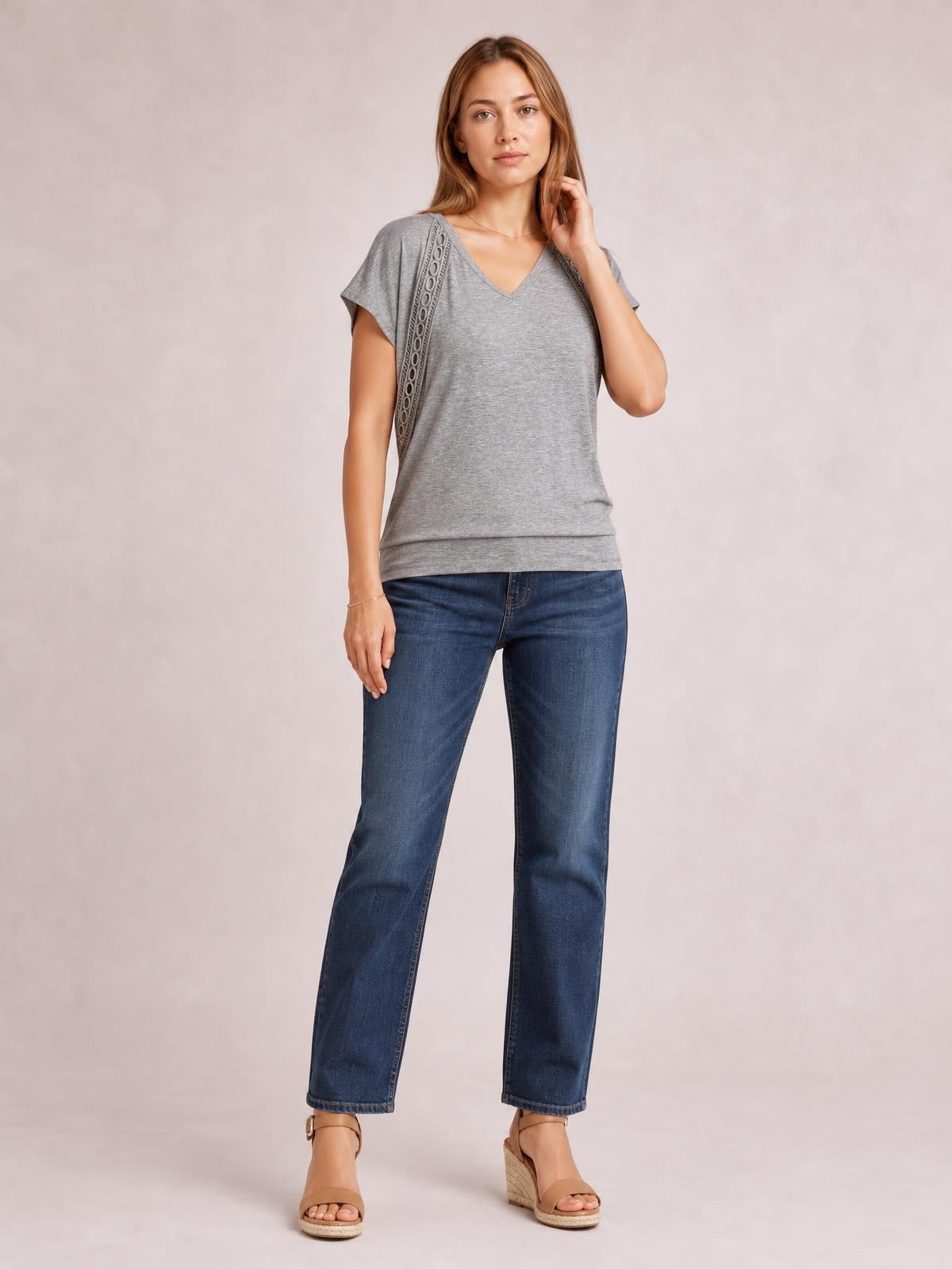 A model poses in a heather gray V-neck short sleeve T-shirt with eyelet shoulder trim, a relaxed straight silhouette, and layered hem.