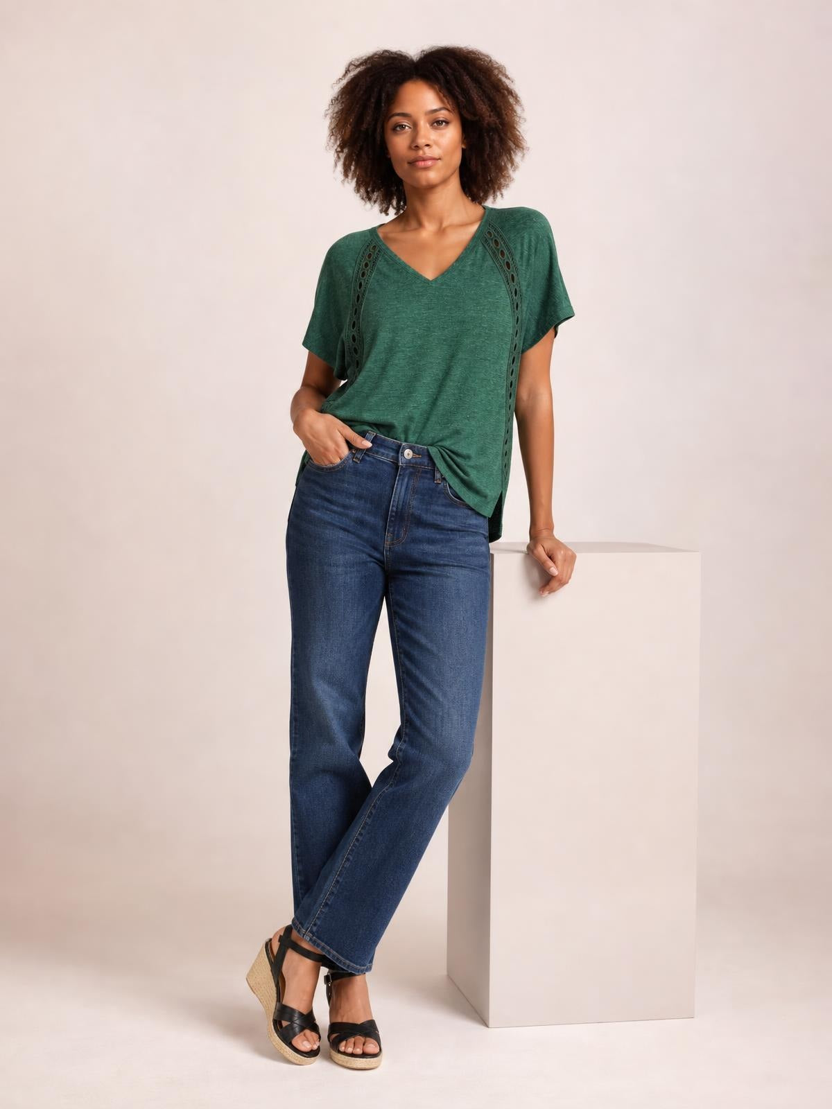 Model wearing a heathered green V-neck short sleeve T-shirt with eyelet side details and a relaxed silhouette, posing with one hand in her jeans pocket.