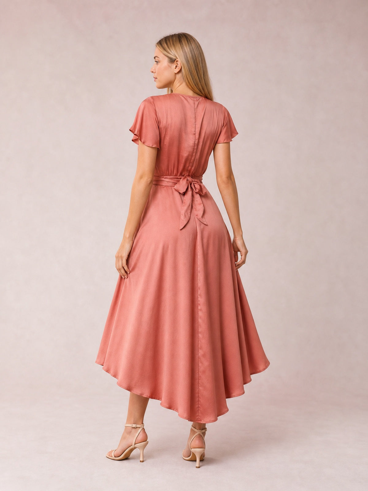 Model shown from the back in a rose pink high-low dress with flutter sleeves, a tied waist, flowing A-line skirt, and visible back seam.