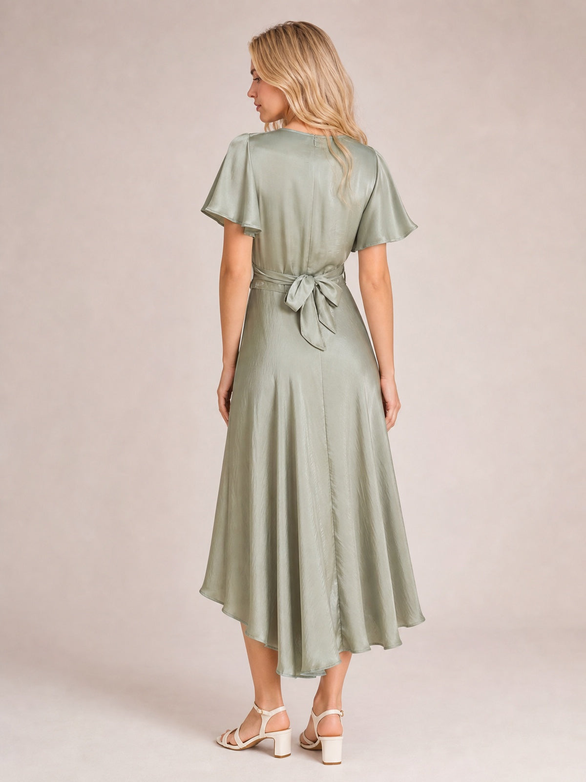 A model wears a sage green high-low dress with flutter sleeves, a flowing A-line skirt, and a tied waist bow, shown from the back in a relaxed pose.
