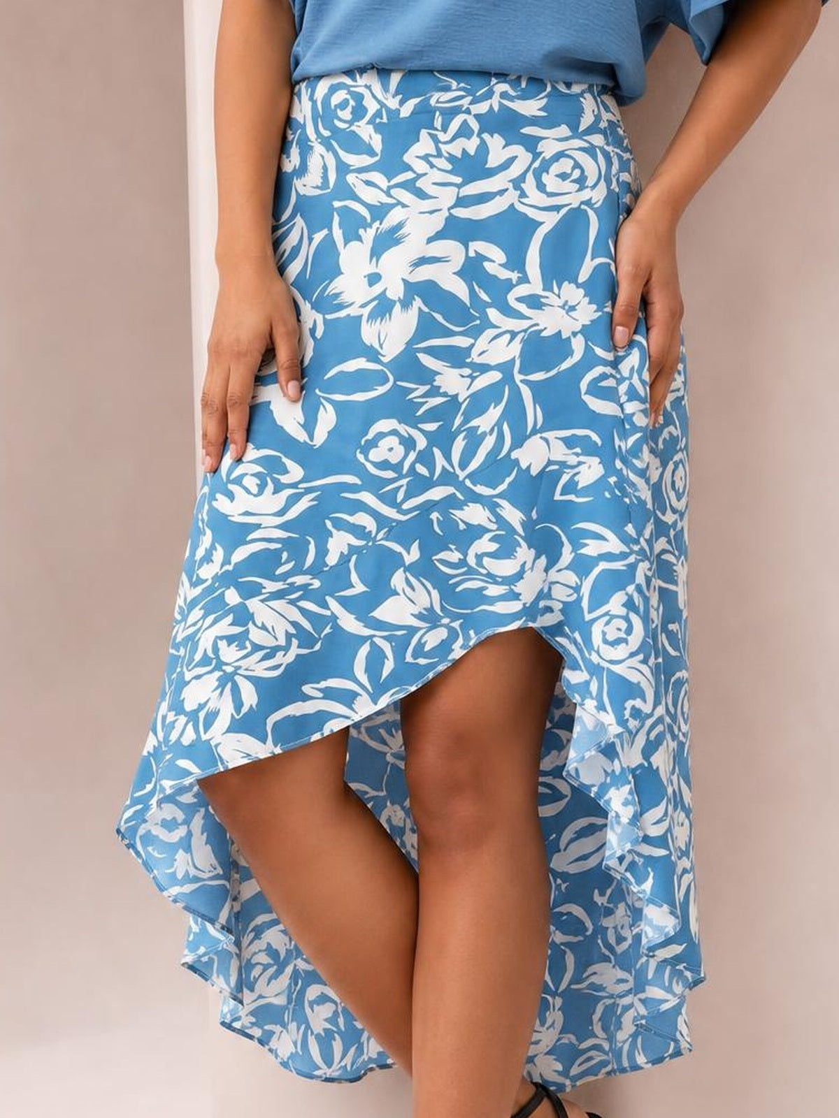 Model wears a blue and white floral printed high-waist skirt with an asymmetrical high-low hem and flowing silhouette, posed with hands at her sides.