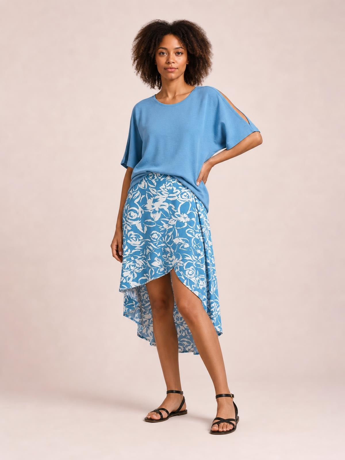 A model poses in a blue and white floral printed high-waist skirt with an asymmetrical high-low hem and flowing silhouette, one hand on her hip.
