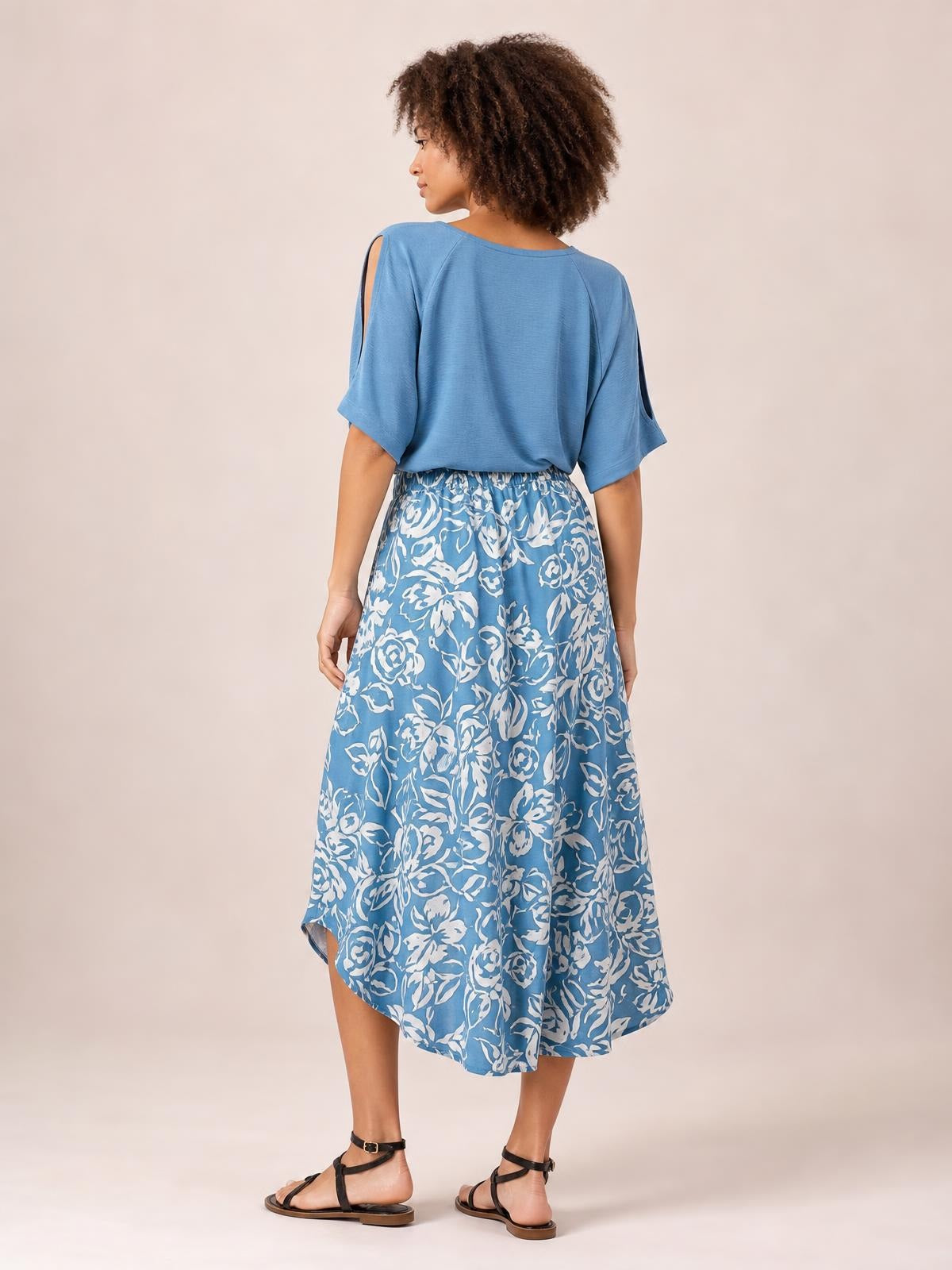 Model shown from behind wearing a blue and white floral printed high-waist skirt with a gathered waistband, flowing silhouette, and asymmetrical high-low hem.