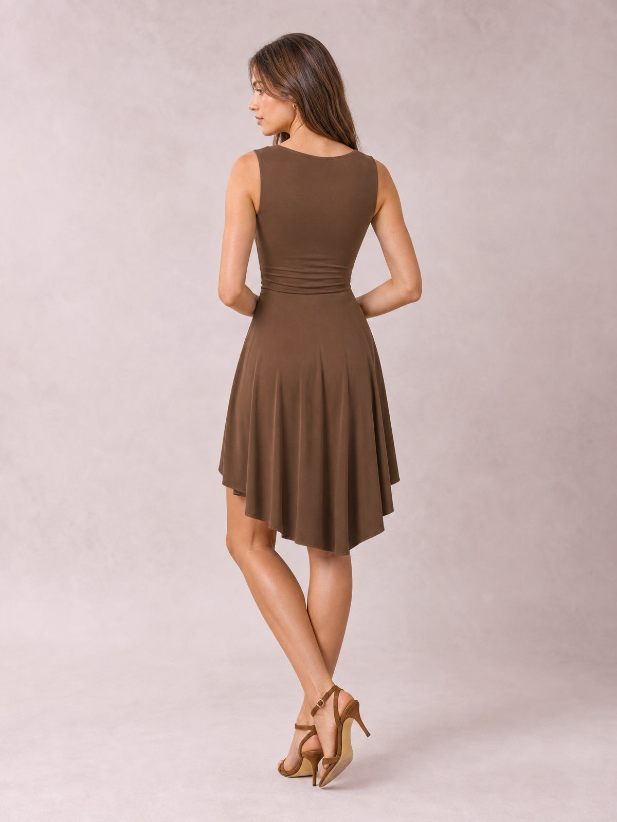 A model poses in a brown sleeveless round-neck mini dress with a fitted waist, softly flared skirt, and flowing high-low hem, viewed from behind.