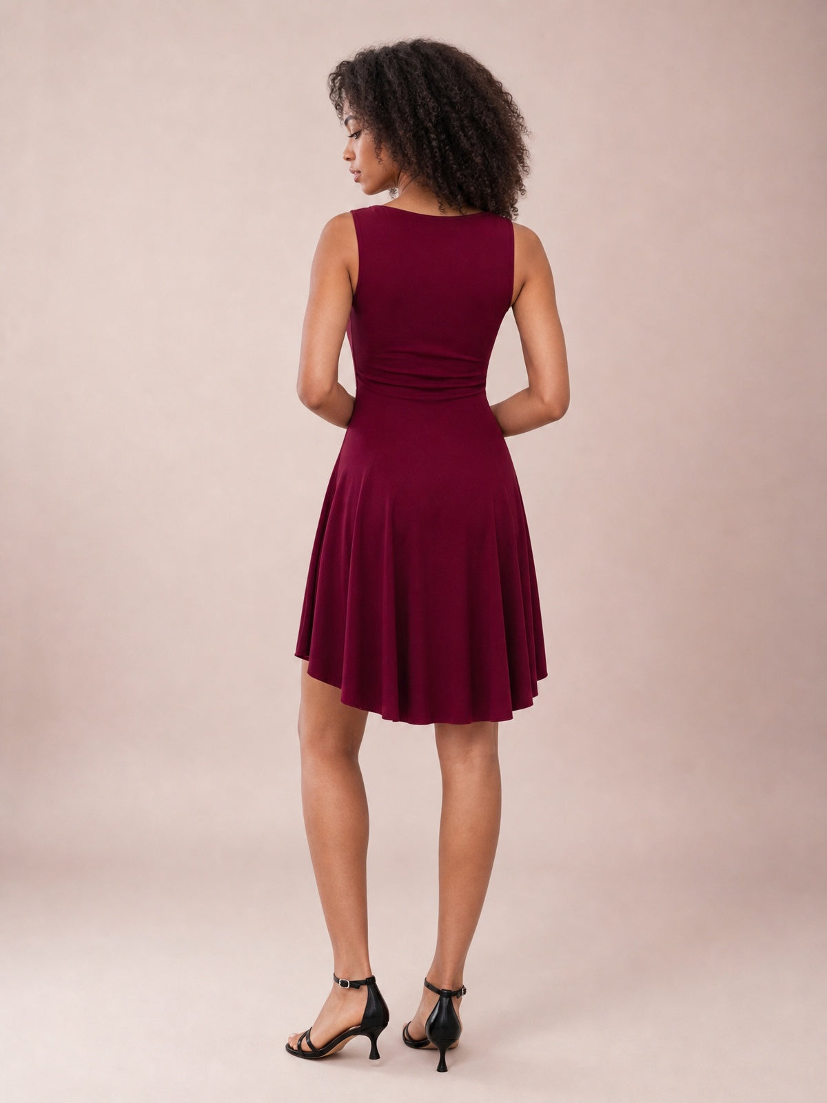 Model poses back-facing in a burgundy high-low round neck sleeveless mini dress with a fitted waist and softly flared skirt.