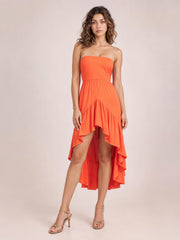 Model stands in a bright orange ruched tube dress with a fitted bodice, gathered waist, and dramatic high-low ruffled hem.