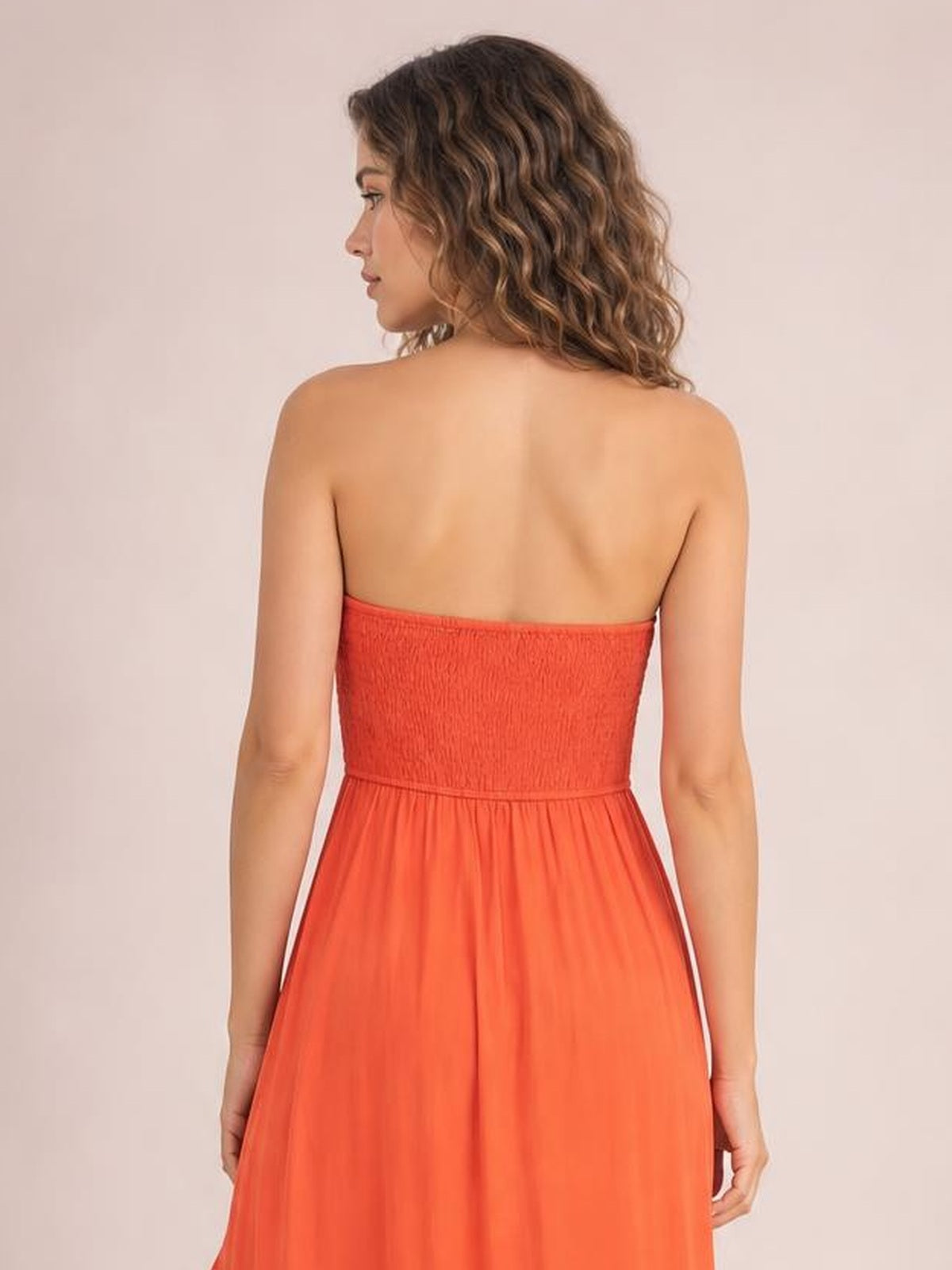 Back view of a model in a coral high-low ruched tube dress with a strapless smocked bodice, defined waist, and flowing skirt silhouette.