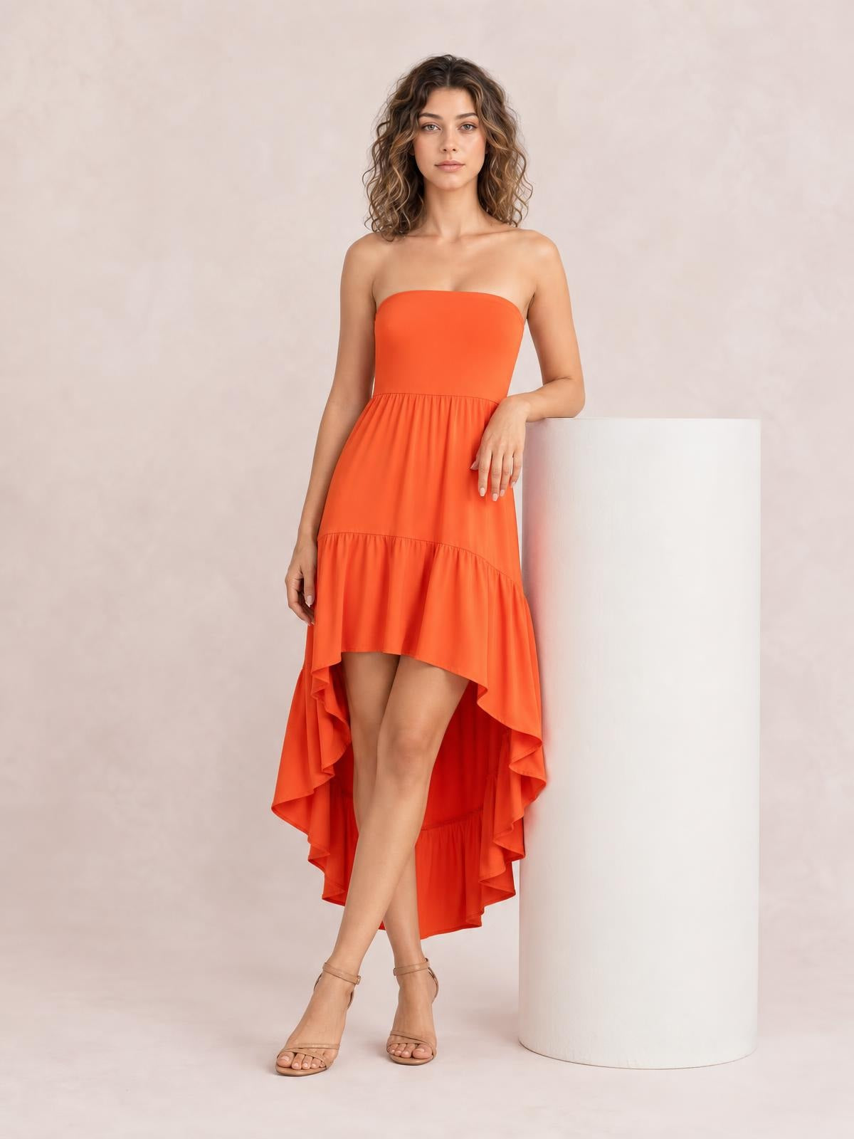 Model in an orange high-low ruched tube dress with a strapless fitted bodice, gathered tiered skirt, and flowing hem, posing with one hand on a pedestal.