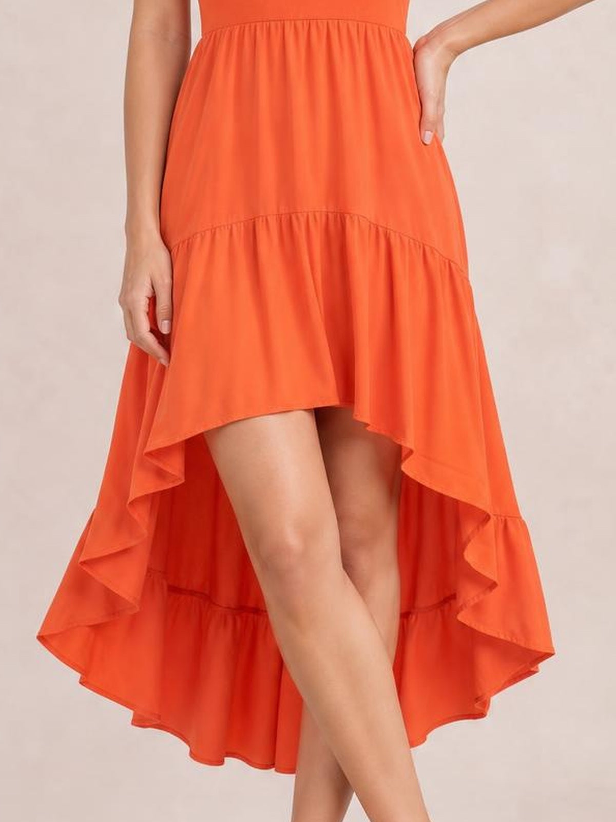 A model poses with crossed legs in a bright orange ruched tube dress featuring a gathered waist, tiered skirt, and dramatic high-low hem.