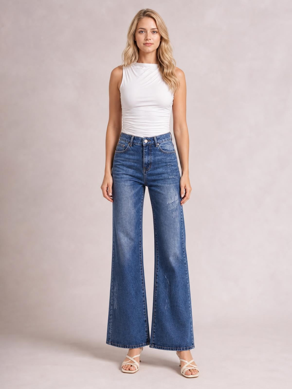 Model stands facing forward in blue high-rise bootcut jeans with a button closure, front pockets, wide flared legs, and subtle distressed detailing.