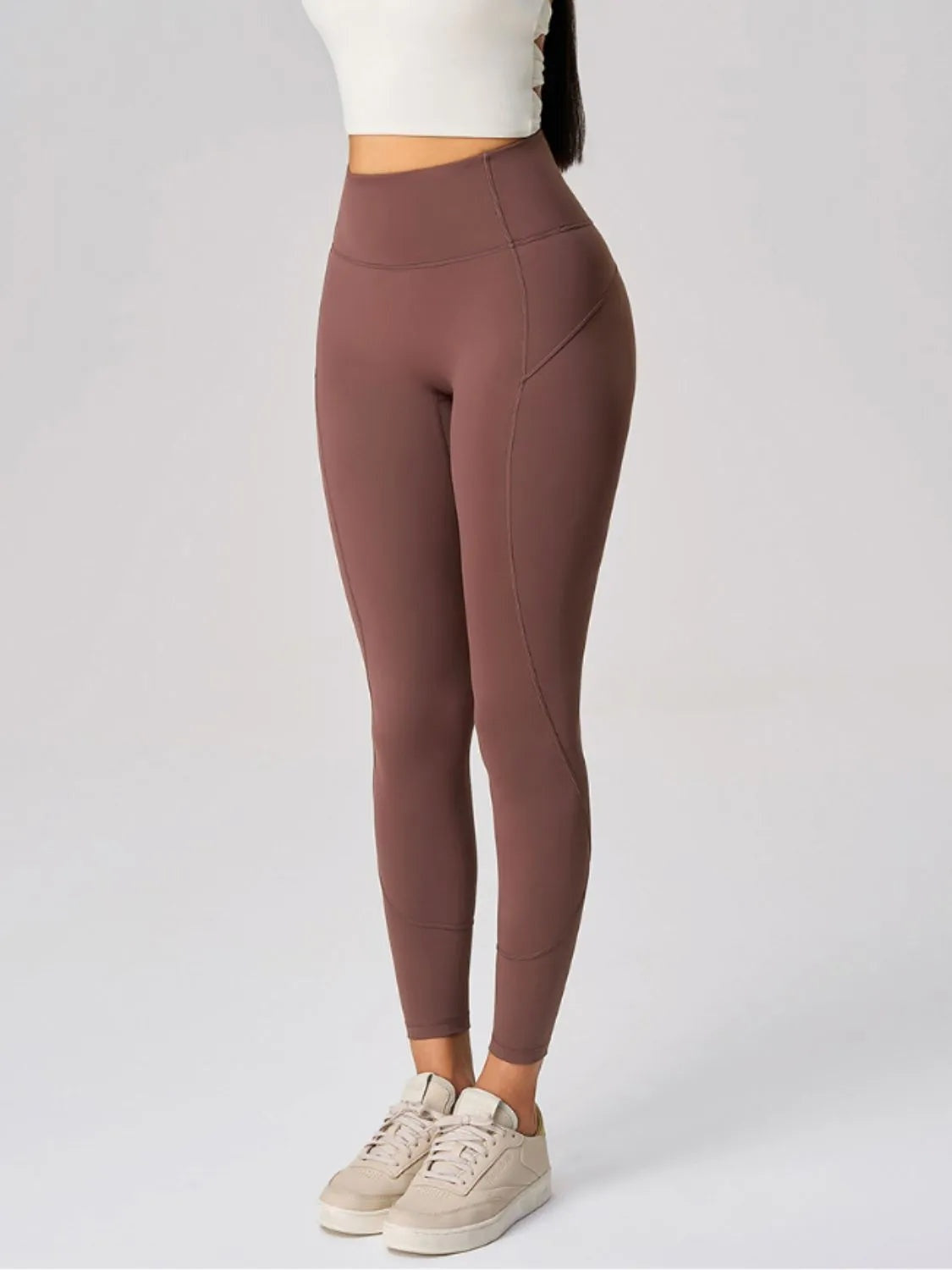 A model stands in brown high-waist active leggings with a fitted ankle-length silhouette, moderate stretch, and visible side pocket and panel seams.