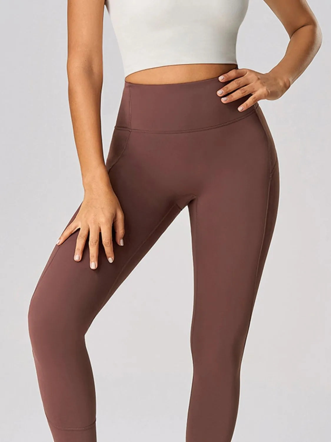 Model poses in brown high-waist active leggings with a wide waistband, slim full-length silhouette, and visible seam detailing.