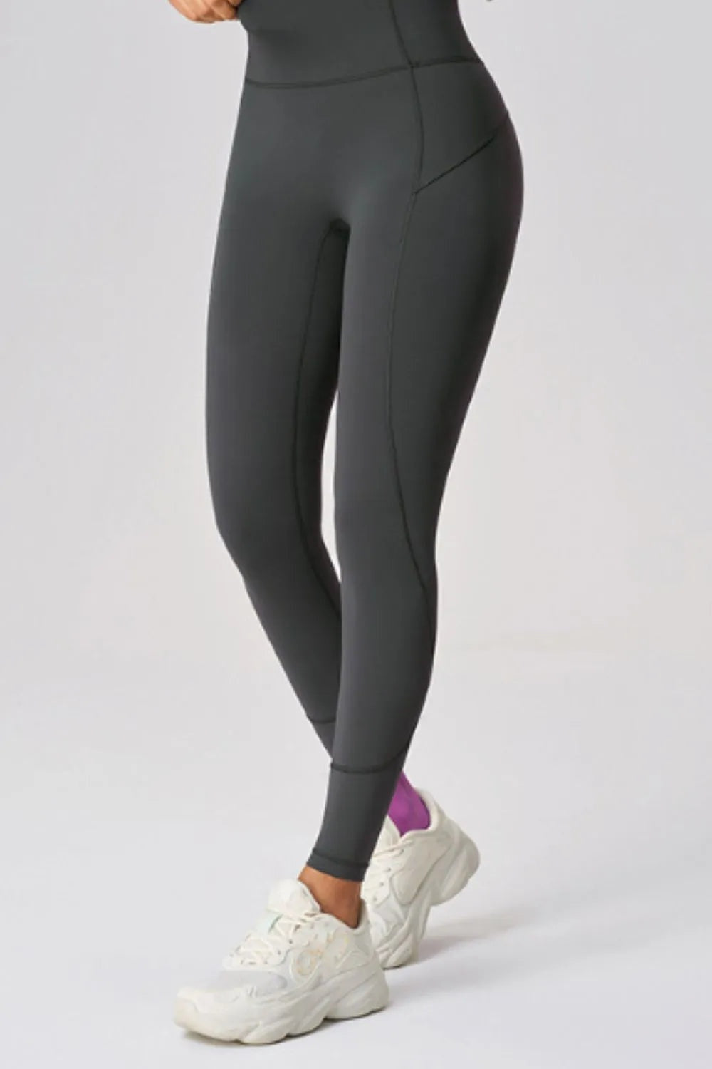 Model wearing black high-waist active leggings with a fitted ankle-length silhouette, contoured seam construction, and one leg crossed in front.