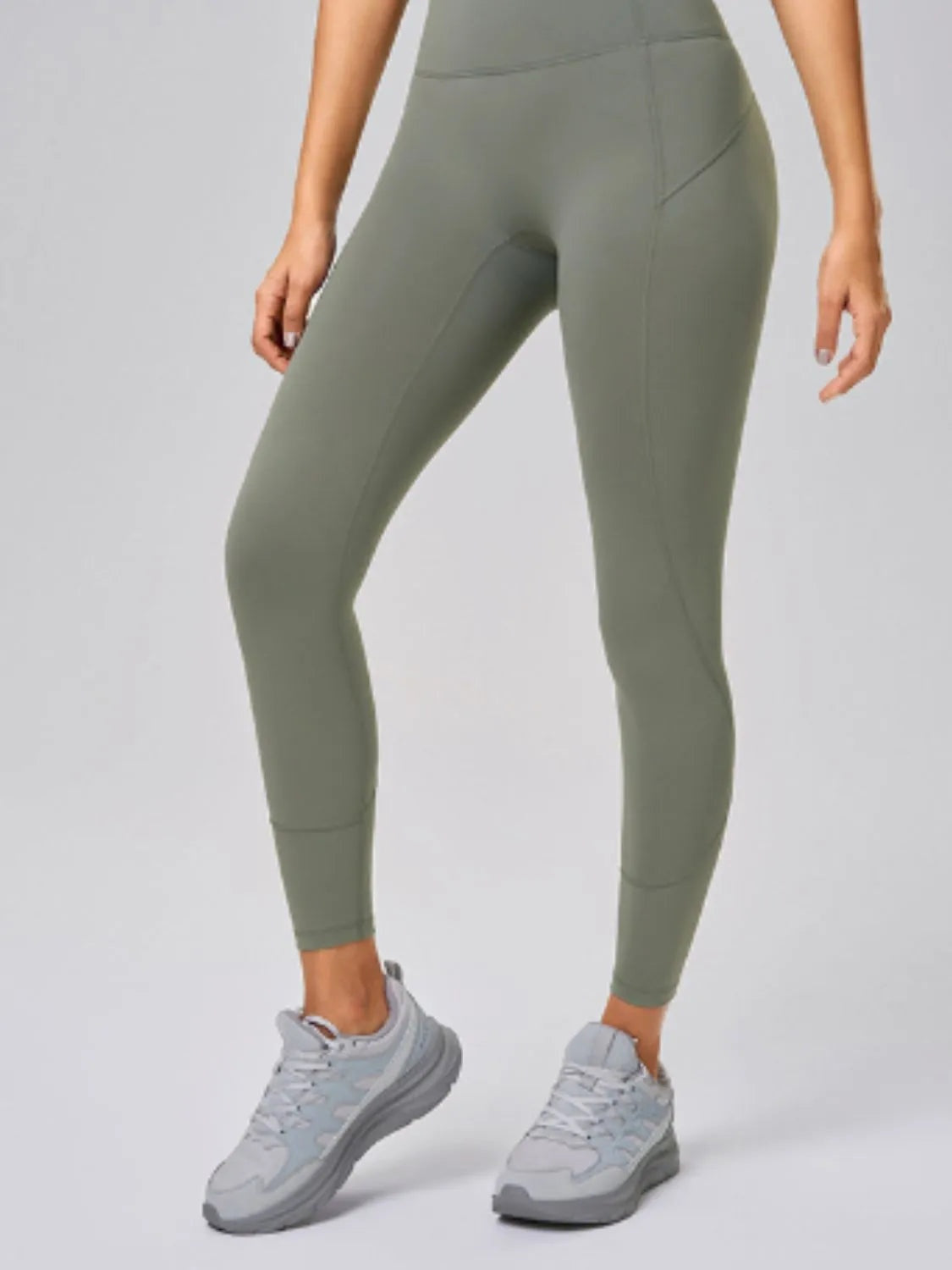 Model wearing olive green high-waist active leggings with a sleek fitted silhouette, ankle-length cuffs, and a visible side pocket, standing with one leg bent.