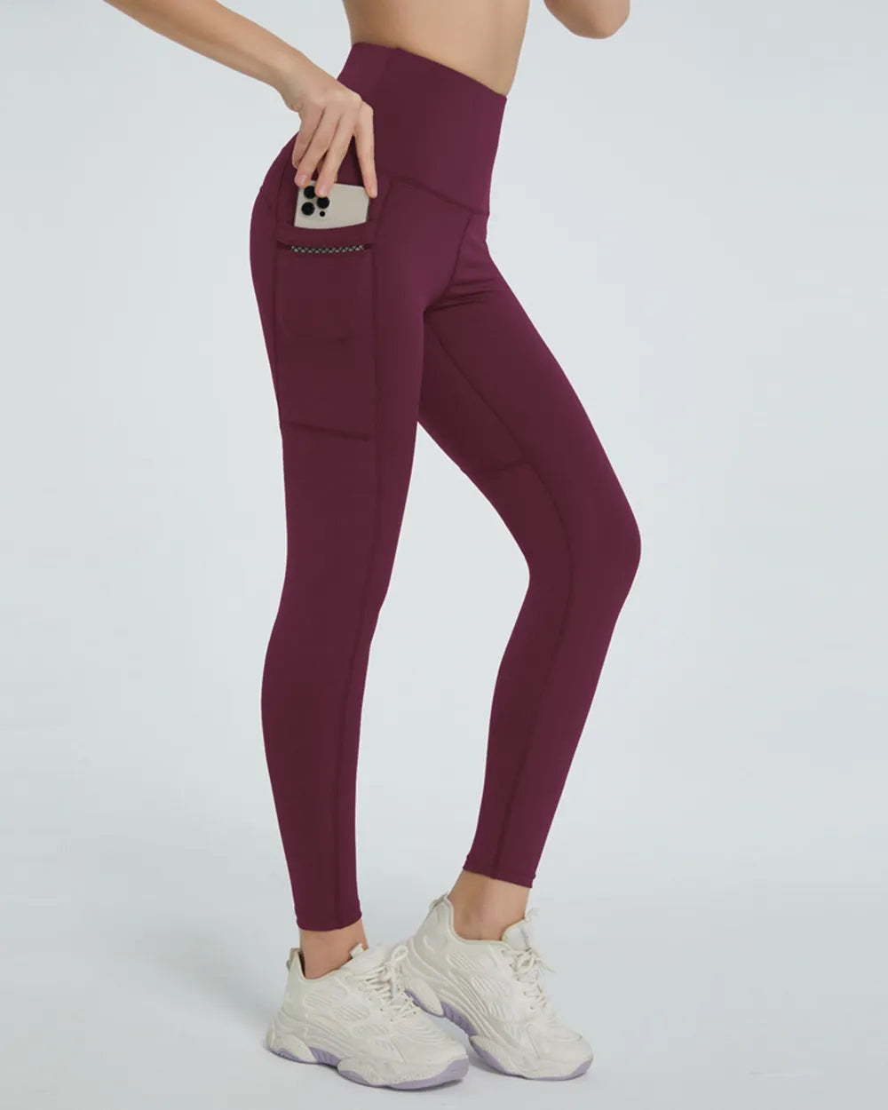 Model poses in burgundy high-waist active leggings with a slim ankle-length silhouette, phone pocket, and visible seam construction.