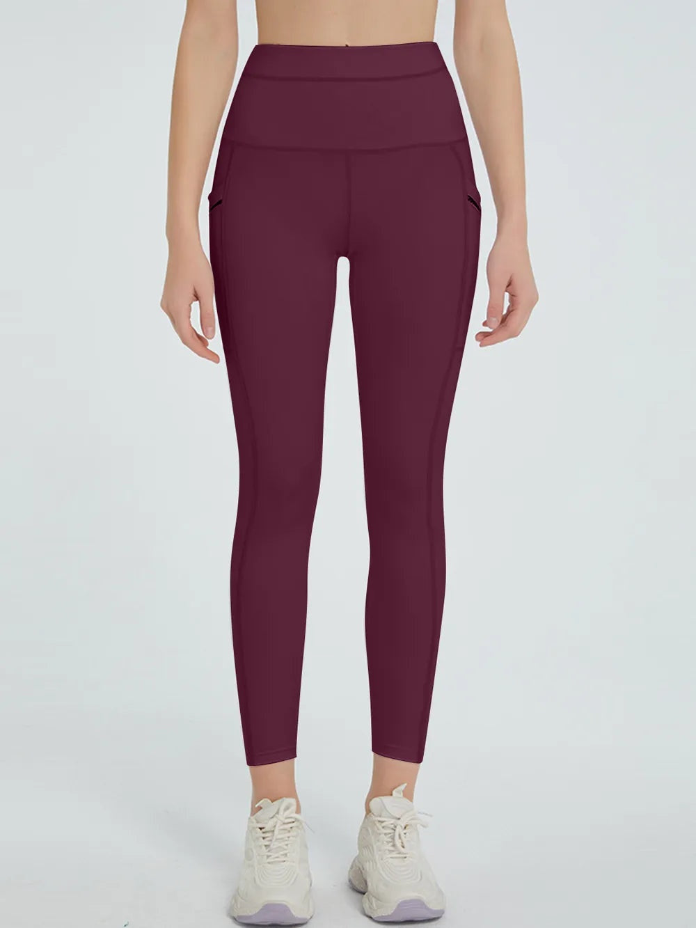 Model standing in burgundy high-waist active leggings with a wide waistband, slim full-length silhouette, and visible side seam pockets.