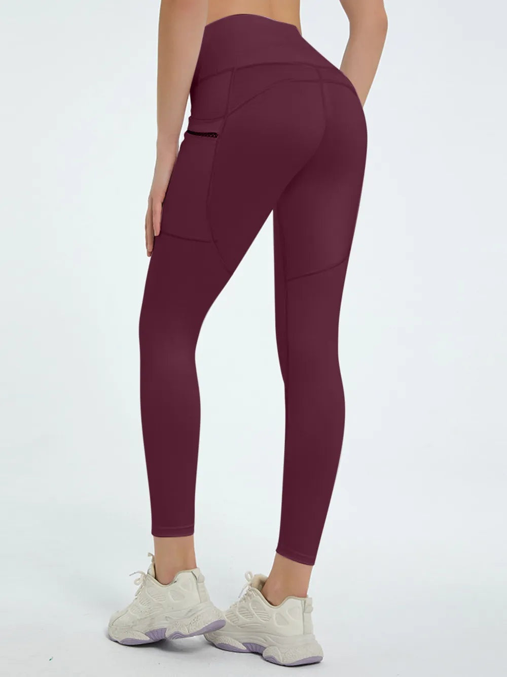 A model stands in a rear three-quarter pose wearing burgundy high waist active leggings with a fitted ankle-length silhouette, curved seams, and a side zip pocket.