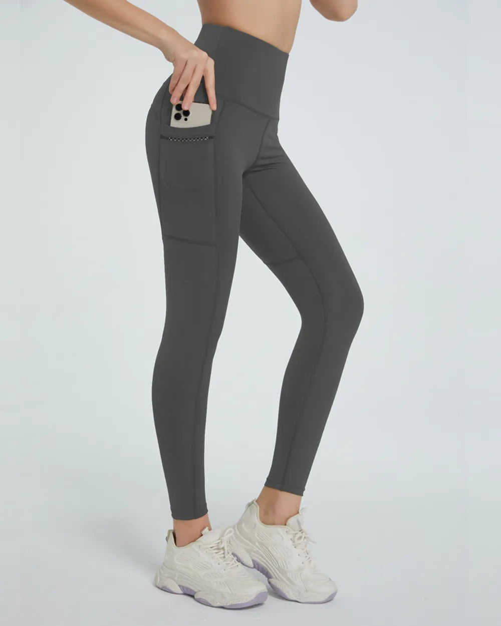 Model wears dark gray high waist active leggings with a slim silhouette, side phone pocket, and visible seam construction while posing in sneakers.