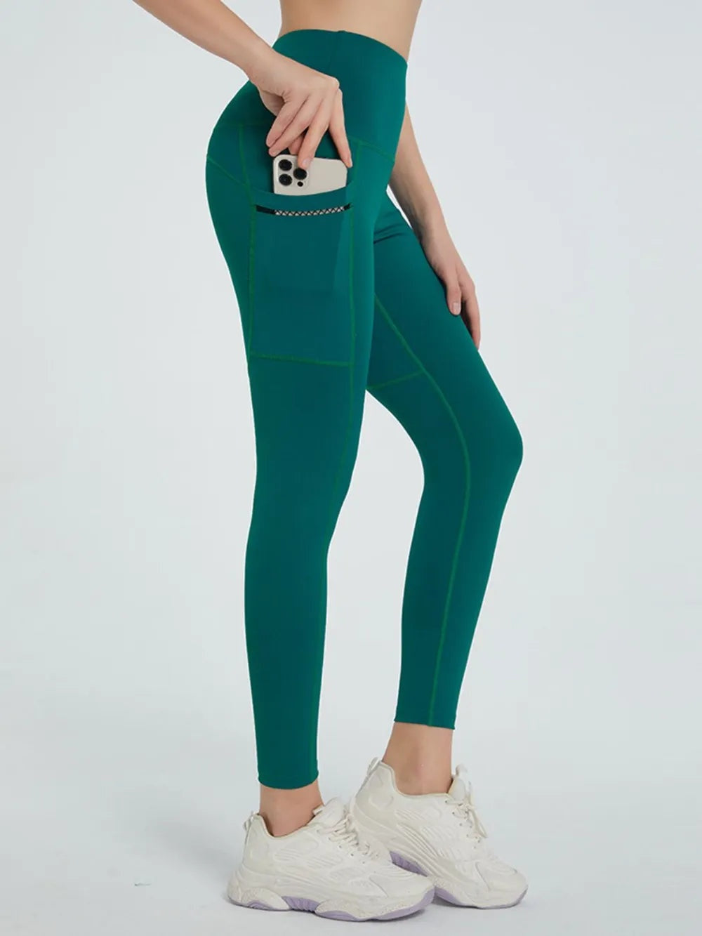 Model poses in dark teal high waist active leggings with a fitted full-length silhouette, visible seam construction, and a side phone pocket.