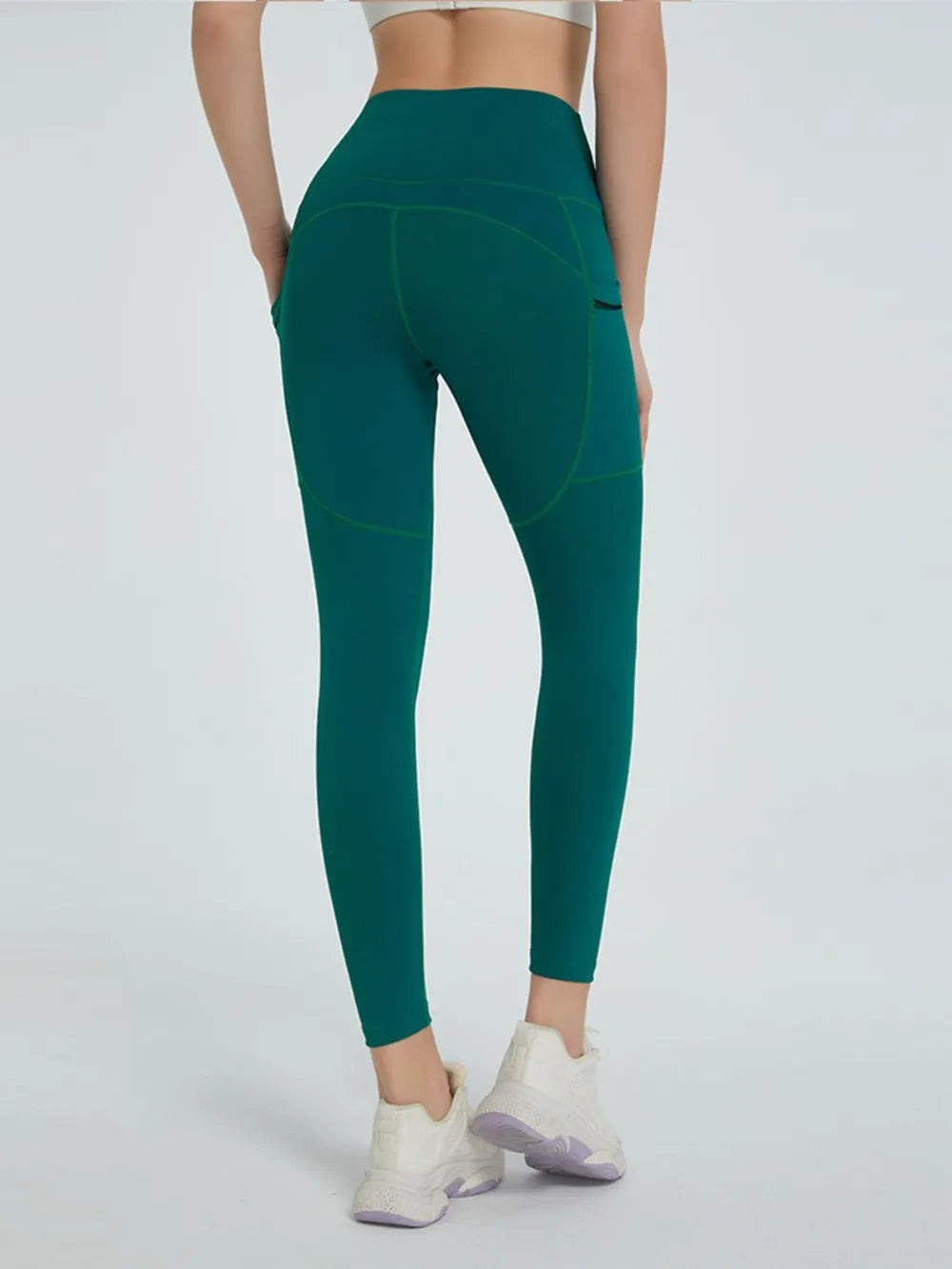 Model standing in dark teal high-waist active leggings with a slim ankle-length silhouette, curved seam detailing, and legs crossed at the ankles.