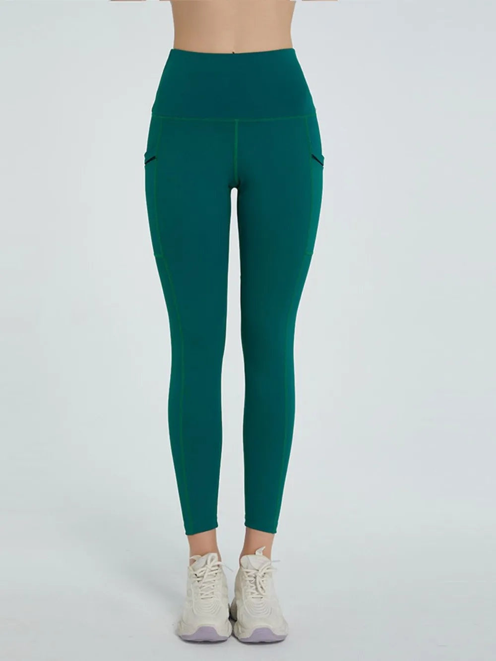 Model standing in dark teal high-waist active leggings with a fitted ankle-length silhouette, stretch construction, and visible side pocket seams.