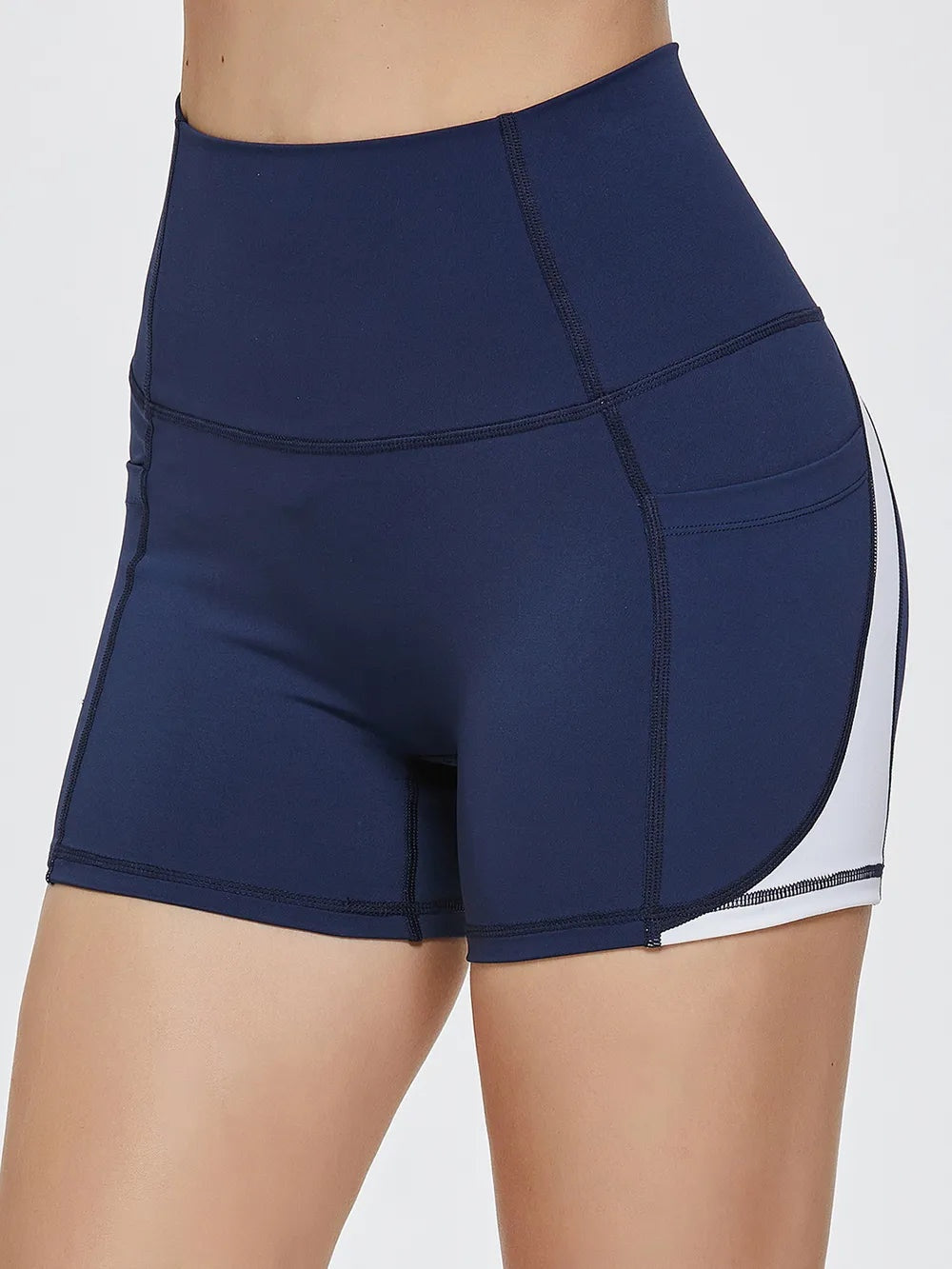 Model wears navy blue high-waist active shorts with a fitted biker-short silhouette, wide waistband, curved panel seams, and contrasting white side trim.