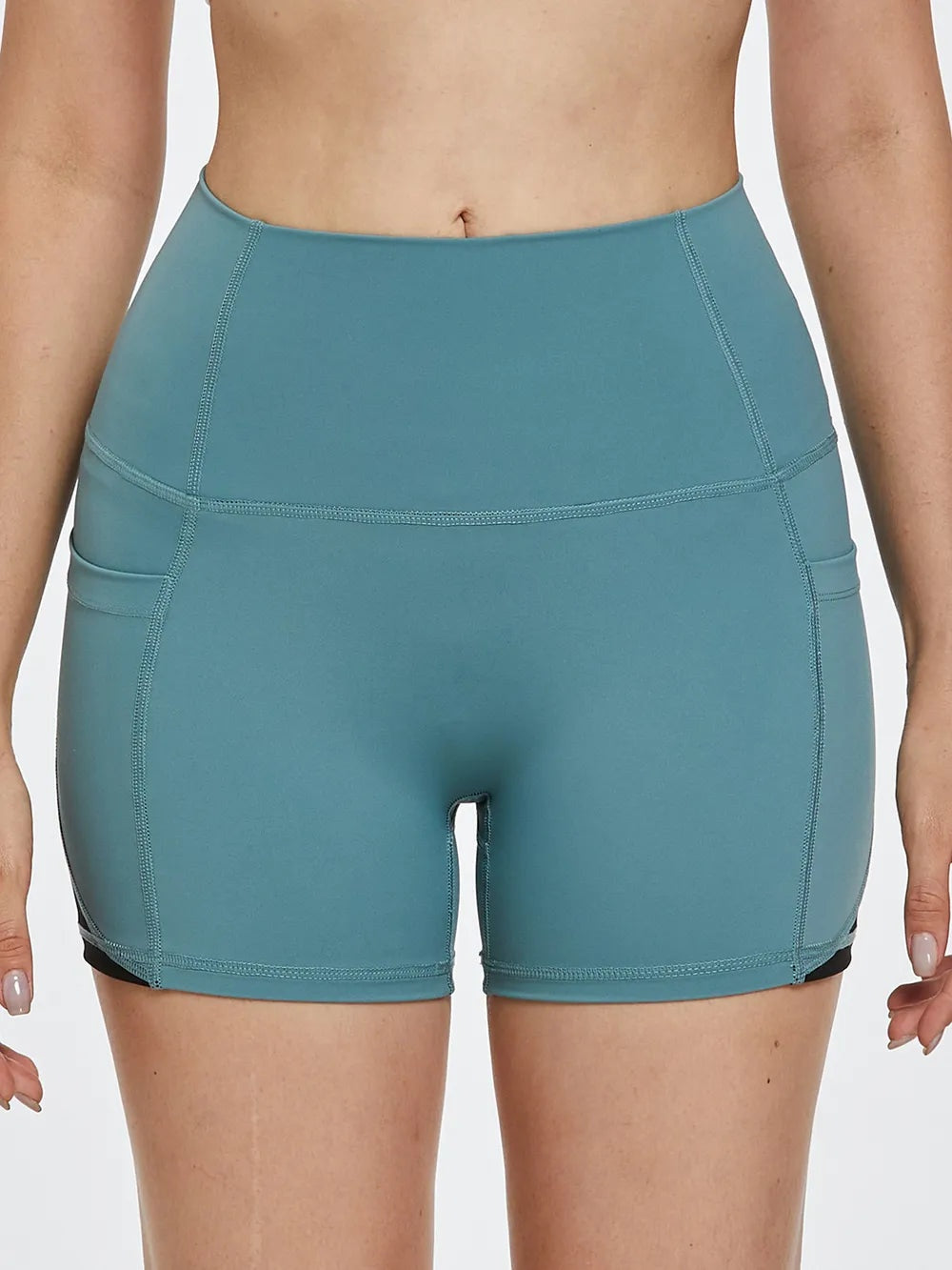 A model wears teal high-waist active shorts in a fitted bike-short silhouette, featuring a wide waistband, curved panel seams, and finished hems.