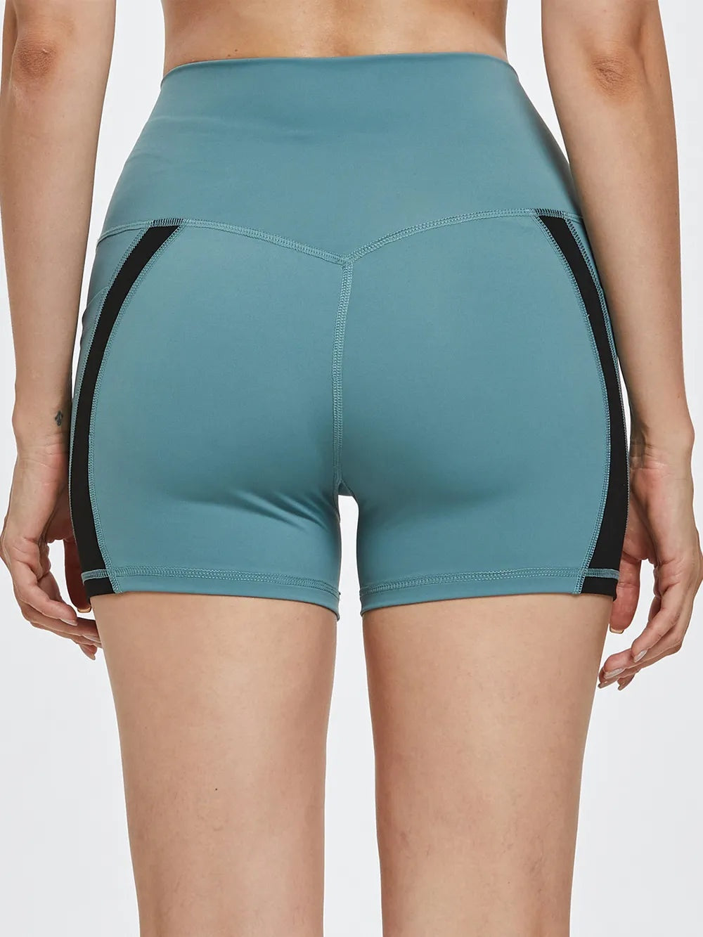 A model stands with hands at her sides wearing teal high-waist active shorts with a close-fitting silhouette, black side panels, and visible contrast seams.