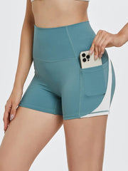 A model wears teal high-waisted active shorts with a fitted bike-short silhouette, visible seam construction, and a side phone pocket while holding a smartphone.