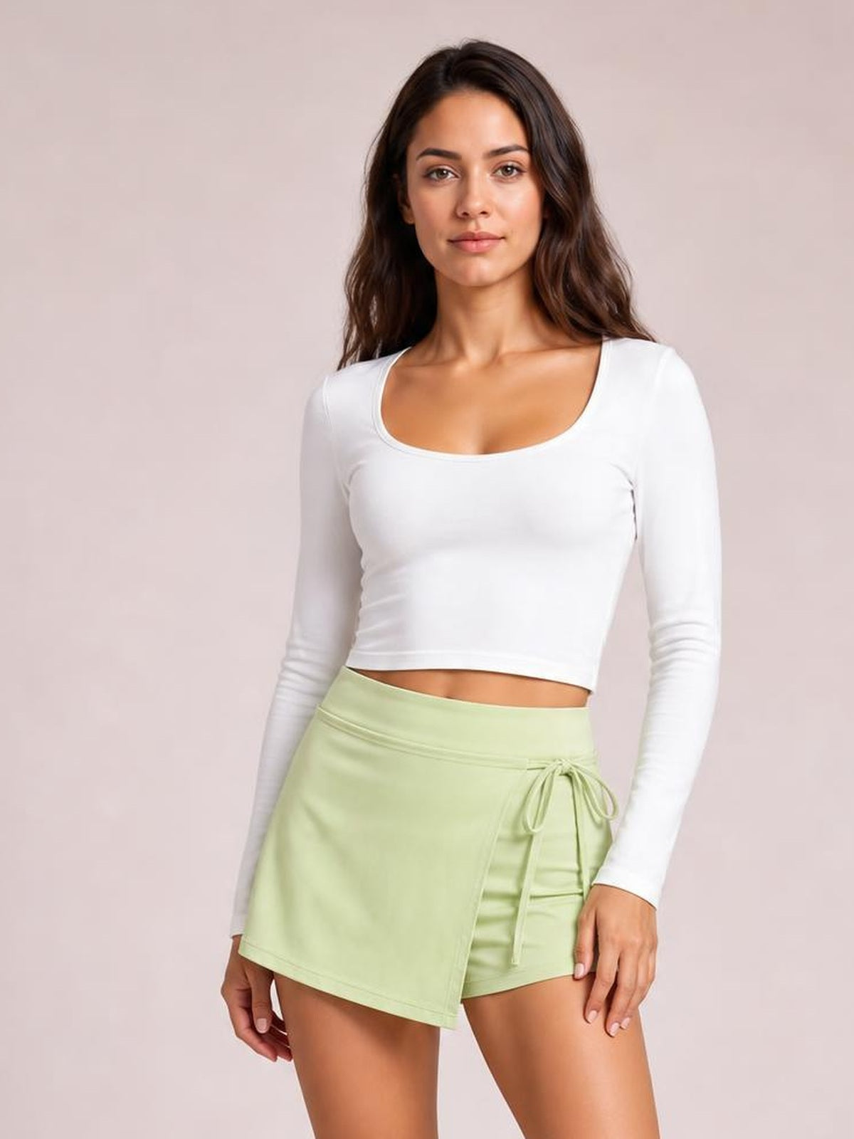 Model poses in a pale green high-waist active skort with an asymmetrical wrap front, built-in shorts, and a tied drawstring waist.