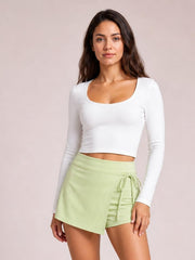 Model poses in a pale green high-waist active skort with an asymmetrical wrap front, built-in shorts, and a tied drawstring waist.
