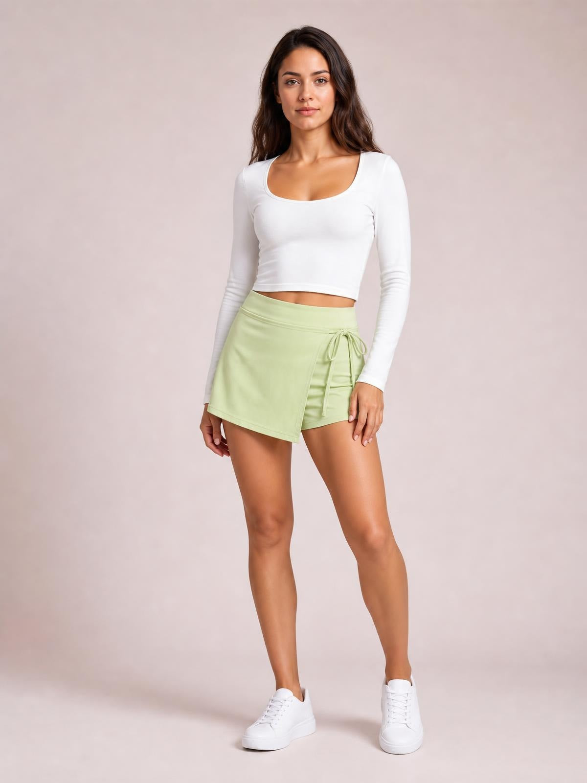 Model stands in a relaxed pose wearing a pale green high-waist active skort with a wrap-front silhouette, tied waist detail, and visible shorts underneath.