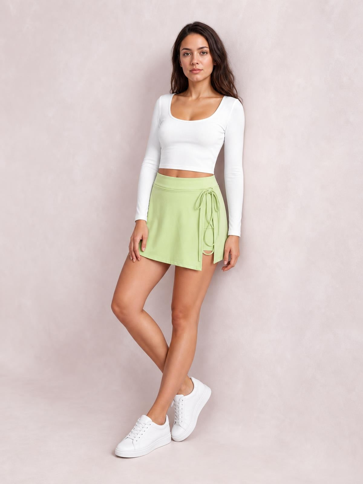 A model poses with crossed legs in a light green high-waist active skort featuring a smooth A-line silhouette, side tie detail, and layered shorts underneath.