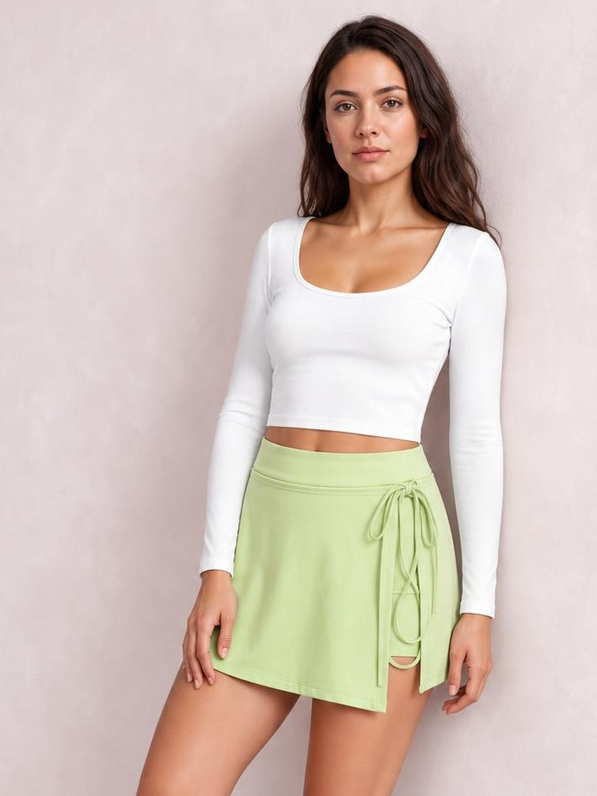A model poses in a light green high waist active skort with a straight silhouette, stretchy construction, and tied side details over fitted shorts.