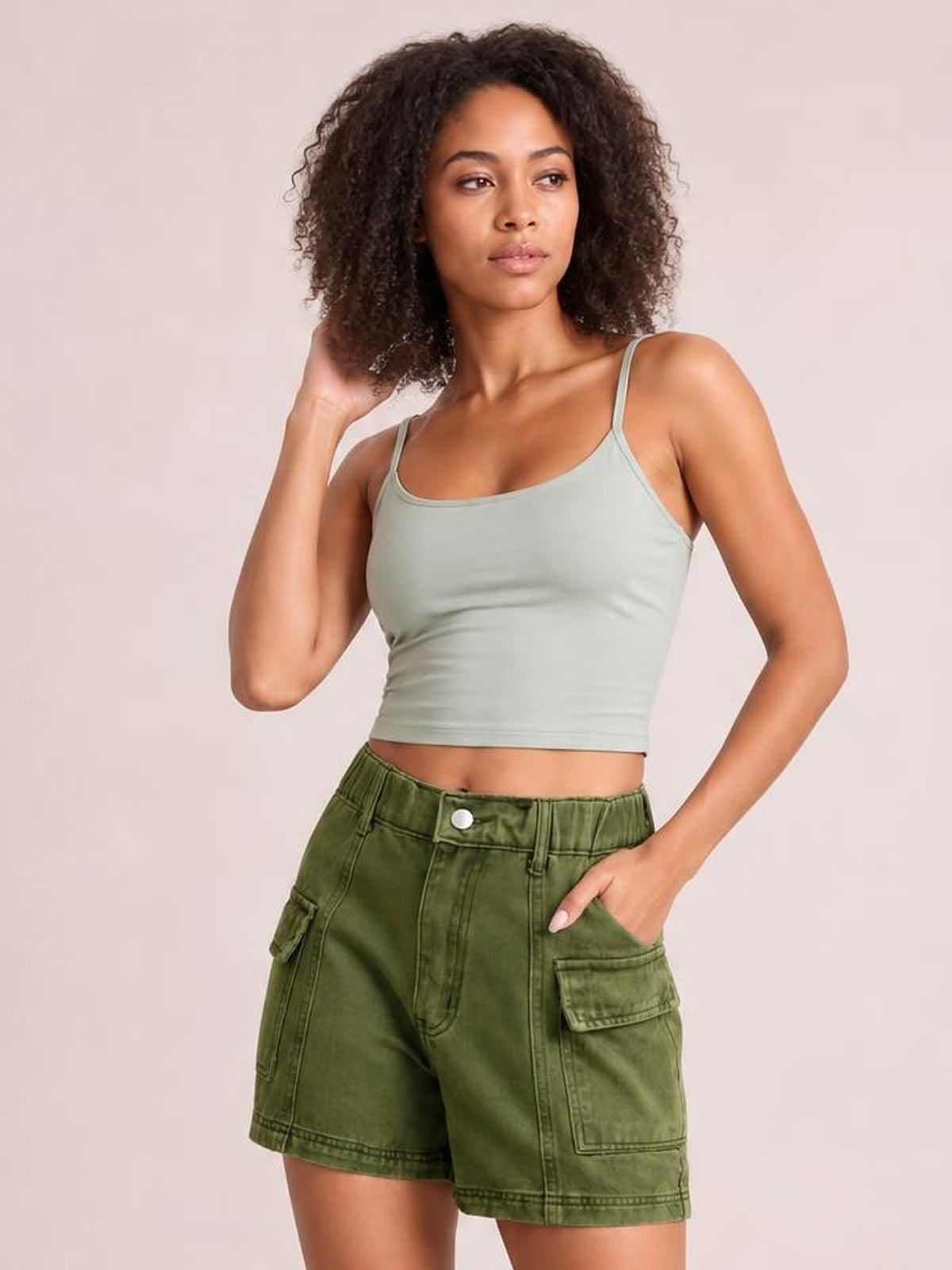 A woman modeling a light sage green ribbed tank top and olive green cargo shorts against a solid pale pink background.