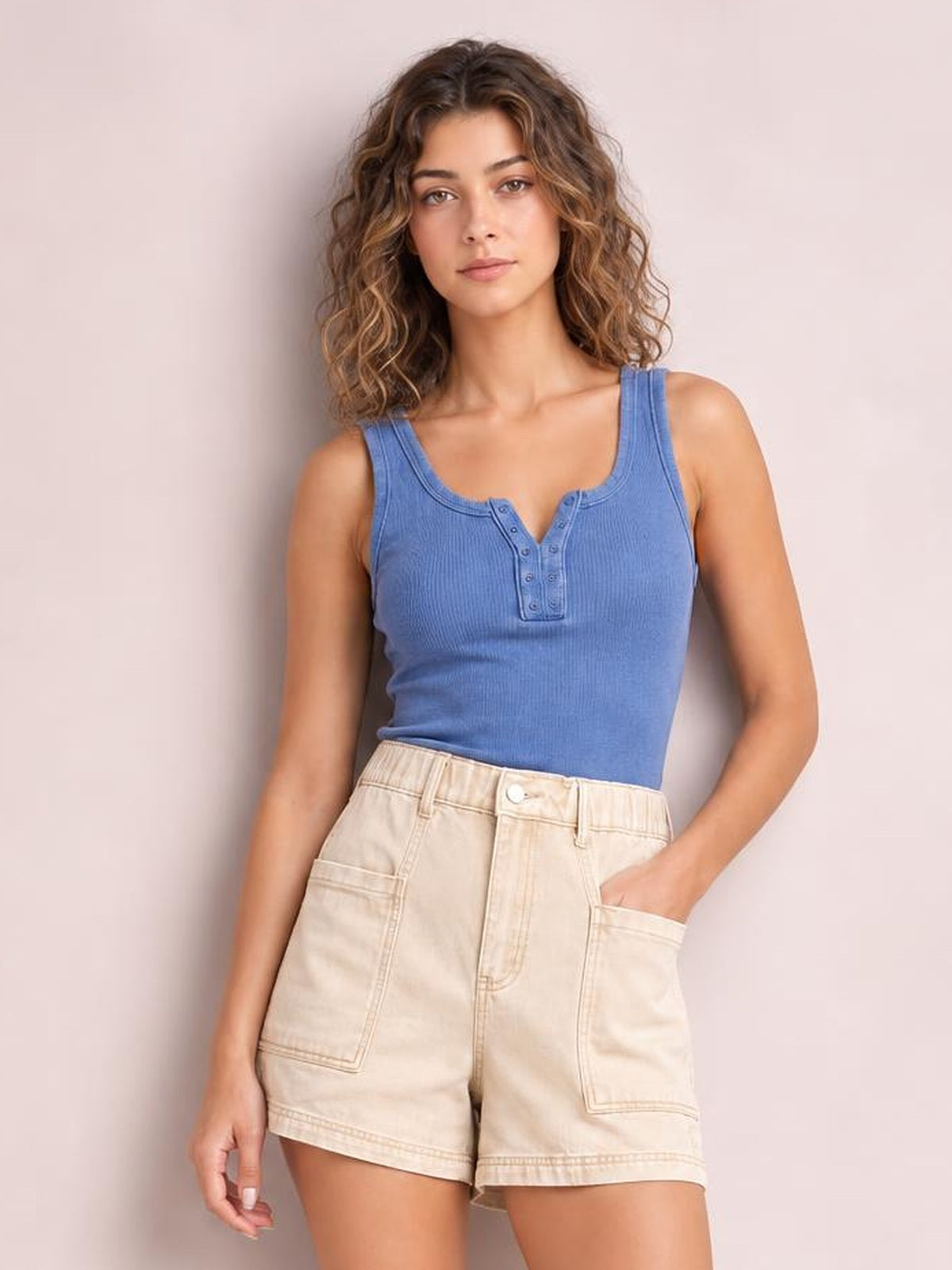 A woman modeling a blue sleeveless henley tank top and cream high-waisted denim shorts against a solid pale pink background.