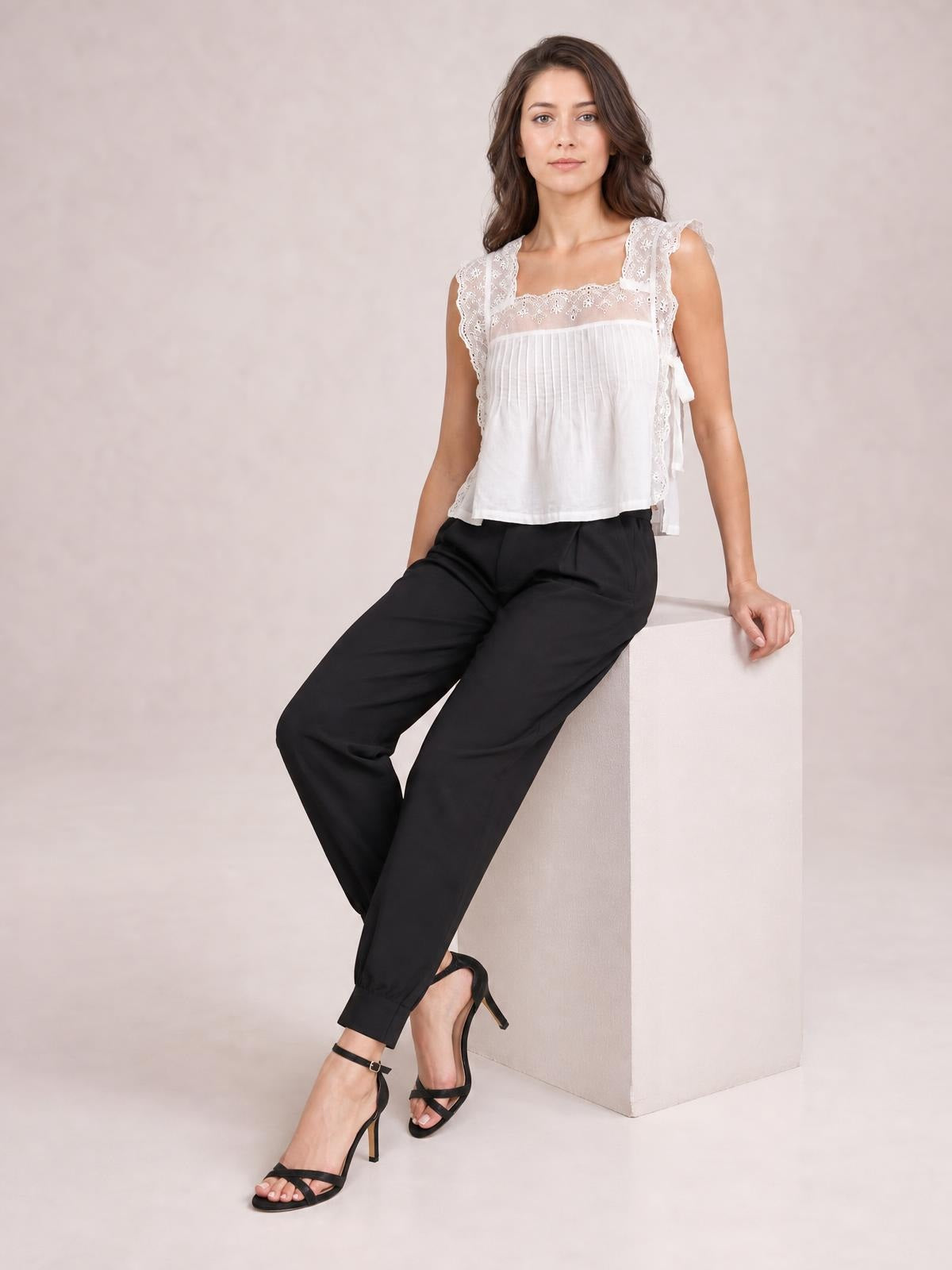 A model sits wearing black high-waisted cropped pants with a tapered silhouette, cuffed hems, and side pockets, posing with one hand tucked inside.