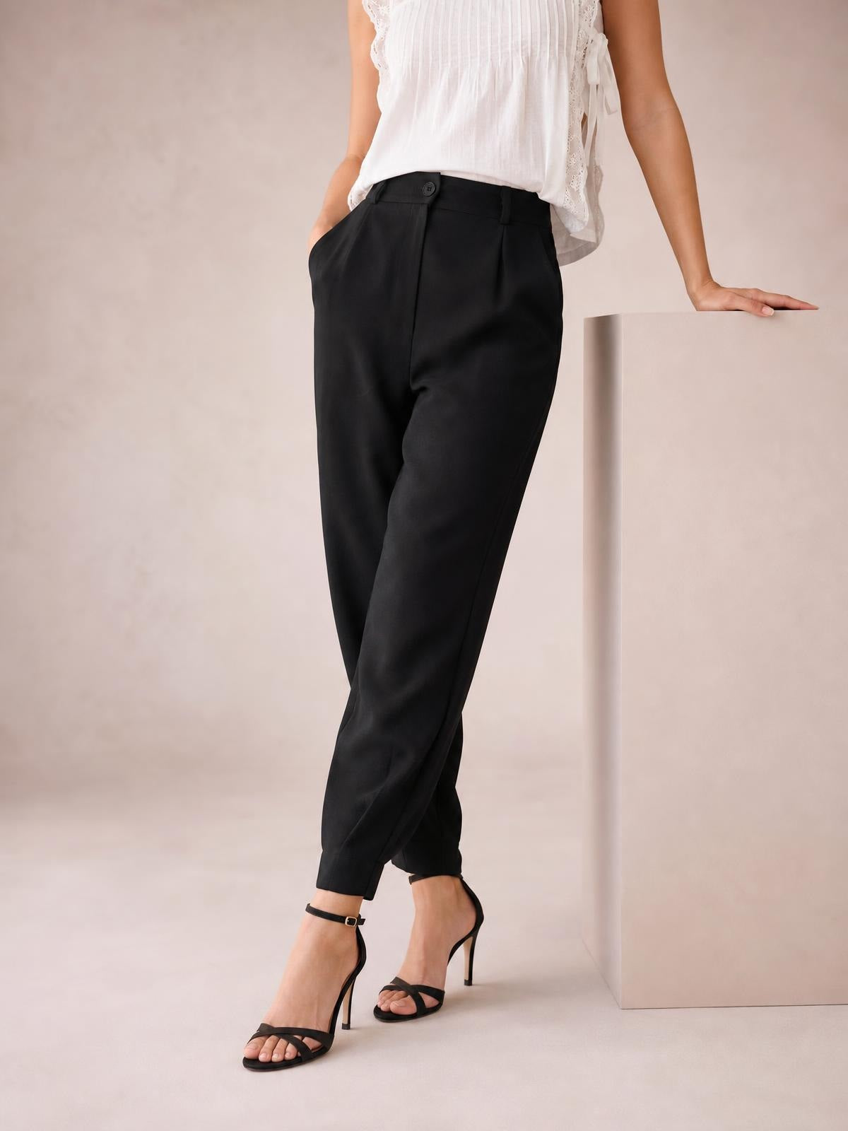 A model poses in black high-waist cropped pants with a tapered silhouette, buttoned waistband, front pleats, and side pockets, one hand tucked inside.